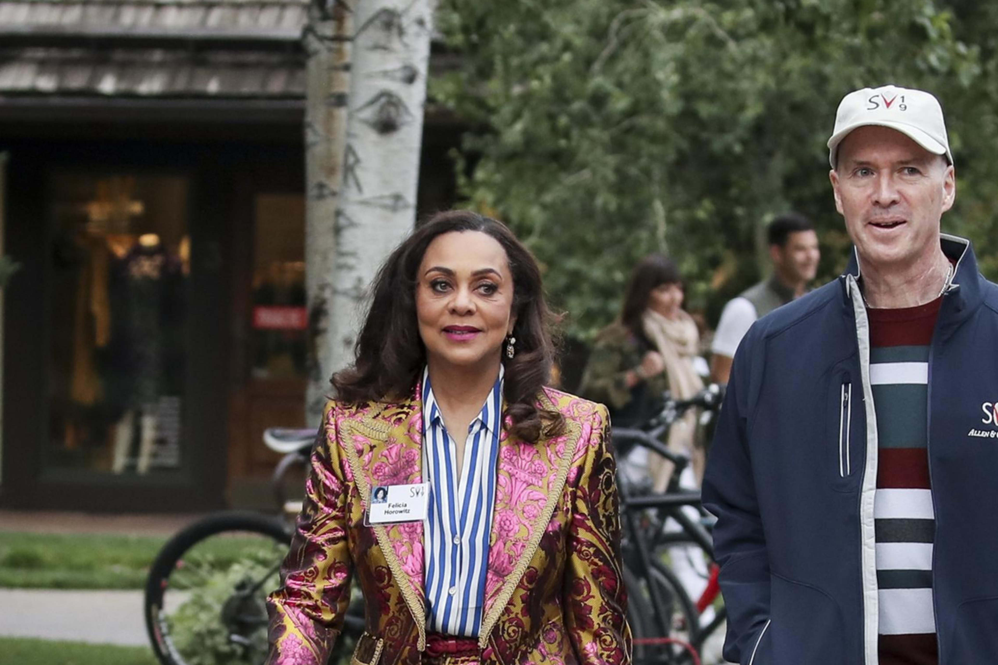 A woman in a vibrant, pink-and-gold baroque-style suit walks alongside a man in casual attire with a striped shirt and a head cap on a paved path with greenery.