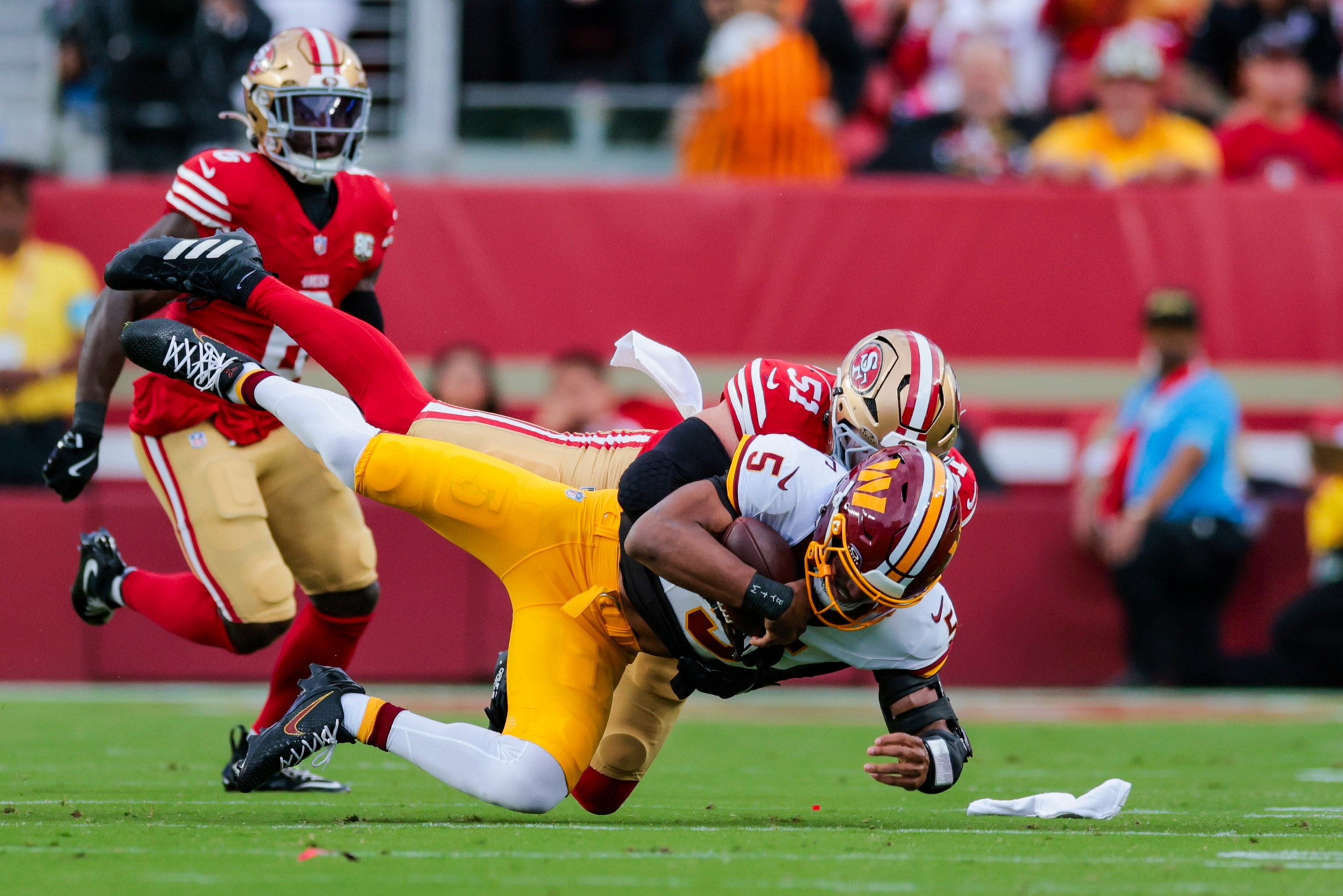 A San Francisco 49ers player tackles a Washington Commanders player who is holding a football, both falling to the grassy field during a game.