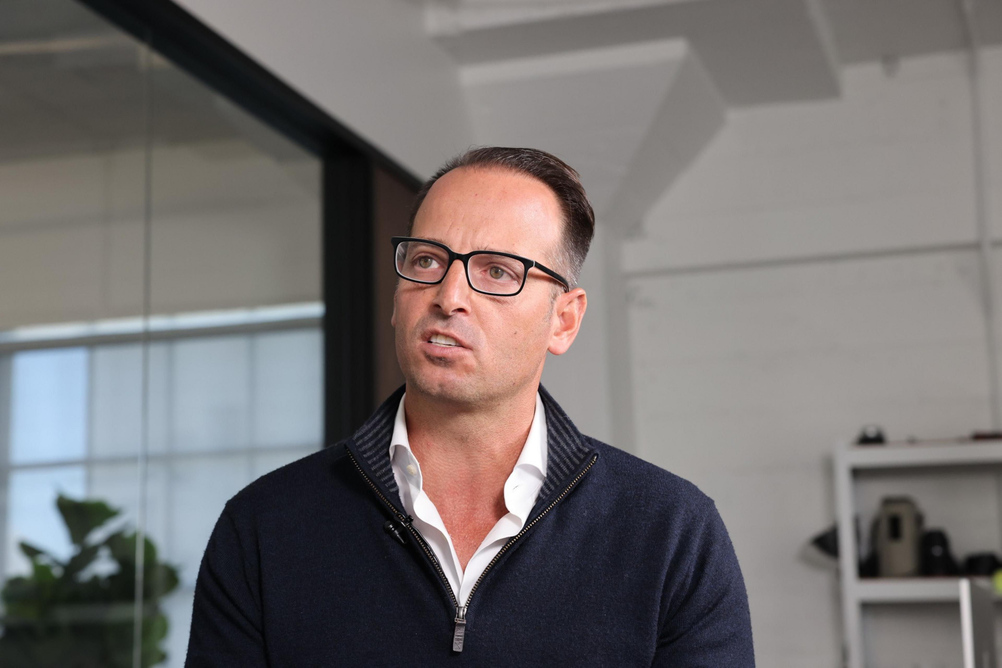A man in glasses, wearing a casual zip-up sweater over a shirt, stands indoors, looking slightly away from the camera.