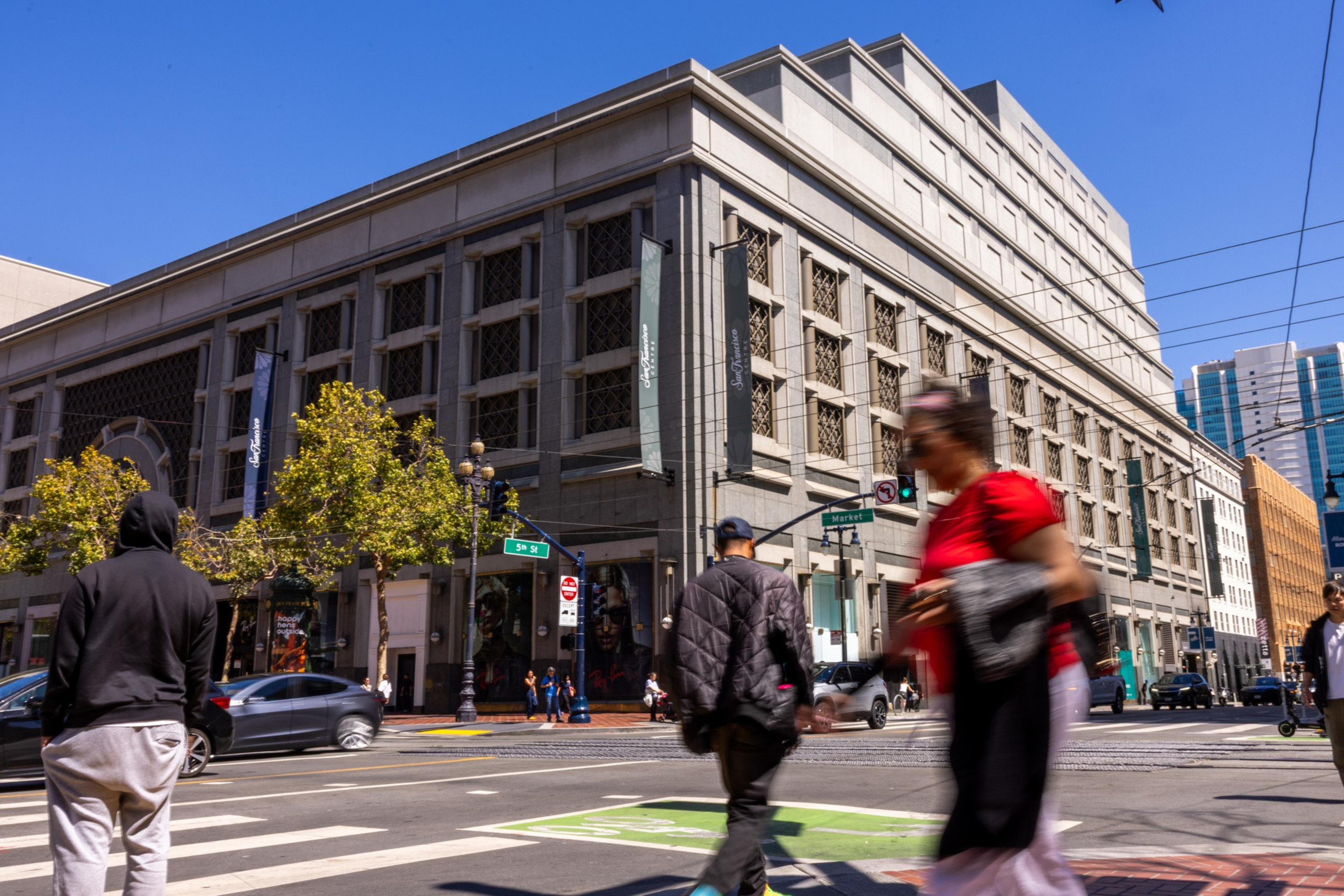People are crossing a city street at an intersection near a large, multi-story building with banners under a clear blue sky.