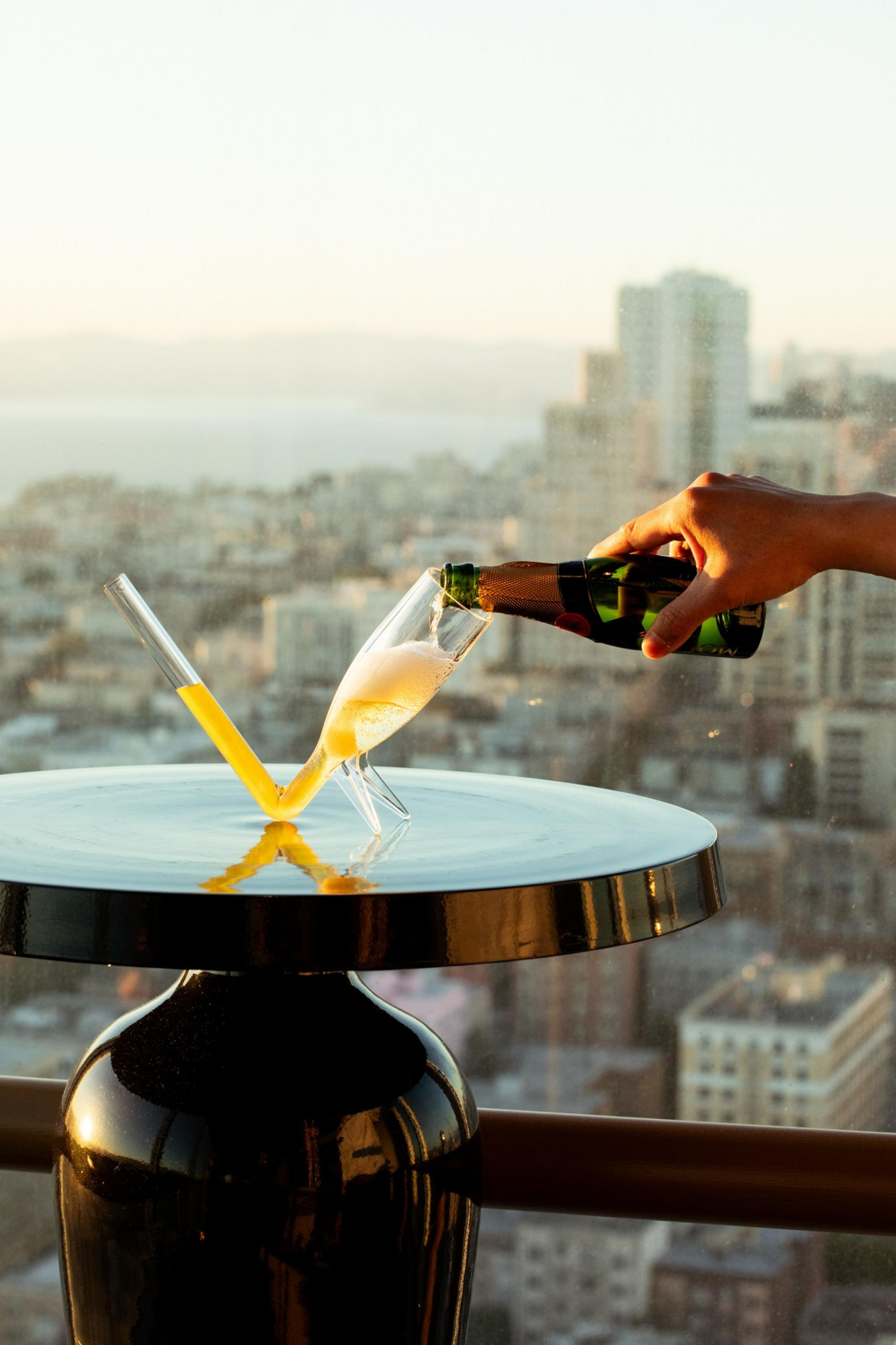 A hand pours champagne into a tilted glass on a shiny round table. Next to it, a flute-like glass with yellow liquid stands amid a cityscape backdrop.