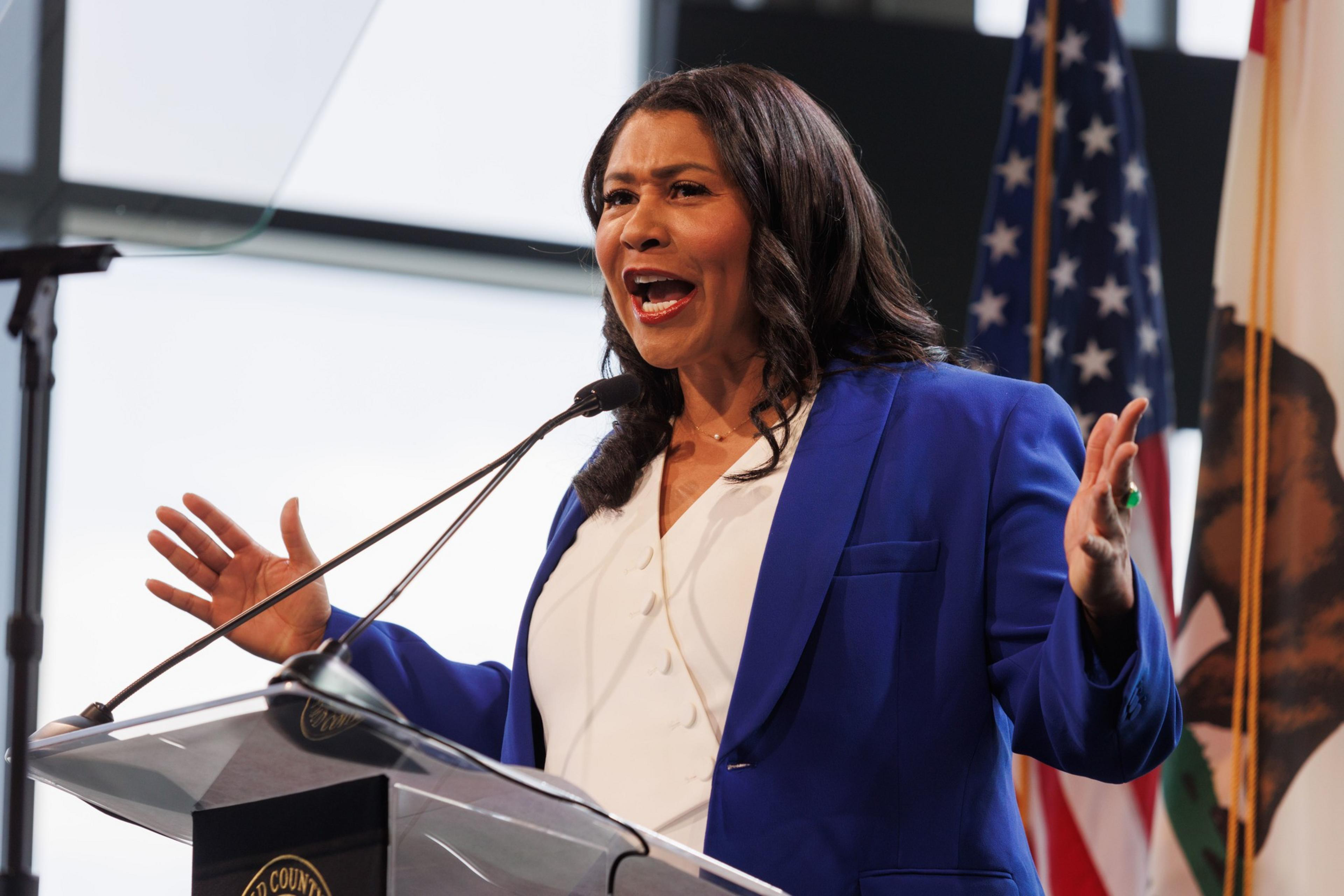 A person wearing a blue blazer speaks animatedly at a lectern with microphones, American flags in the background.