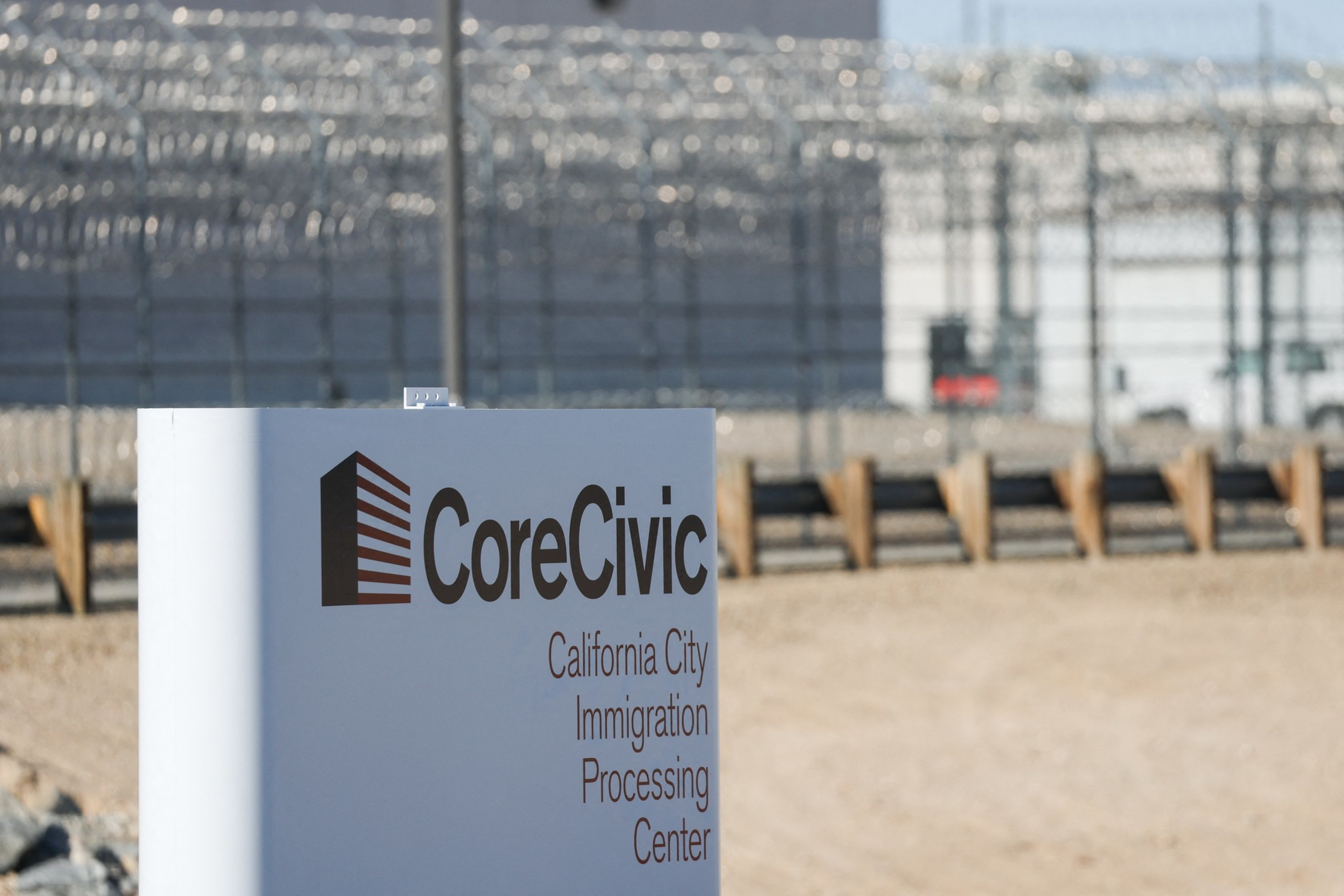 A sign reads “CoreCivic California City Immigration Processing Center” with a barbed wire fence and security structures in the background.