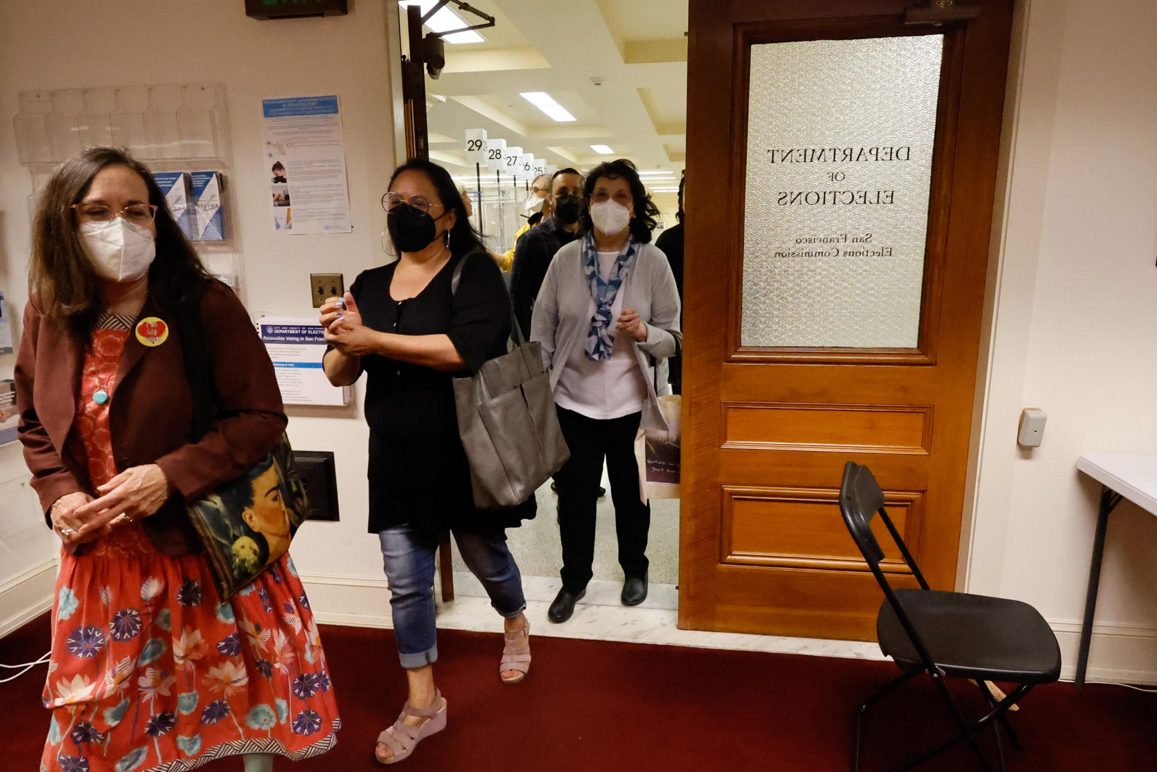 Several people wearing masks walk through a doorway labeled "Department of Elections" inside a building.