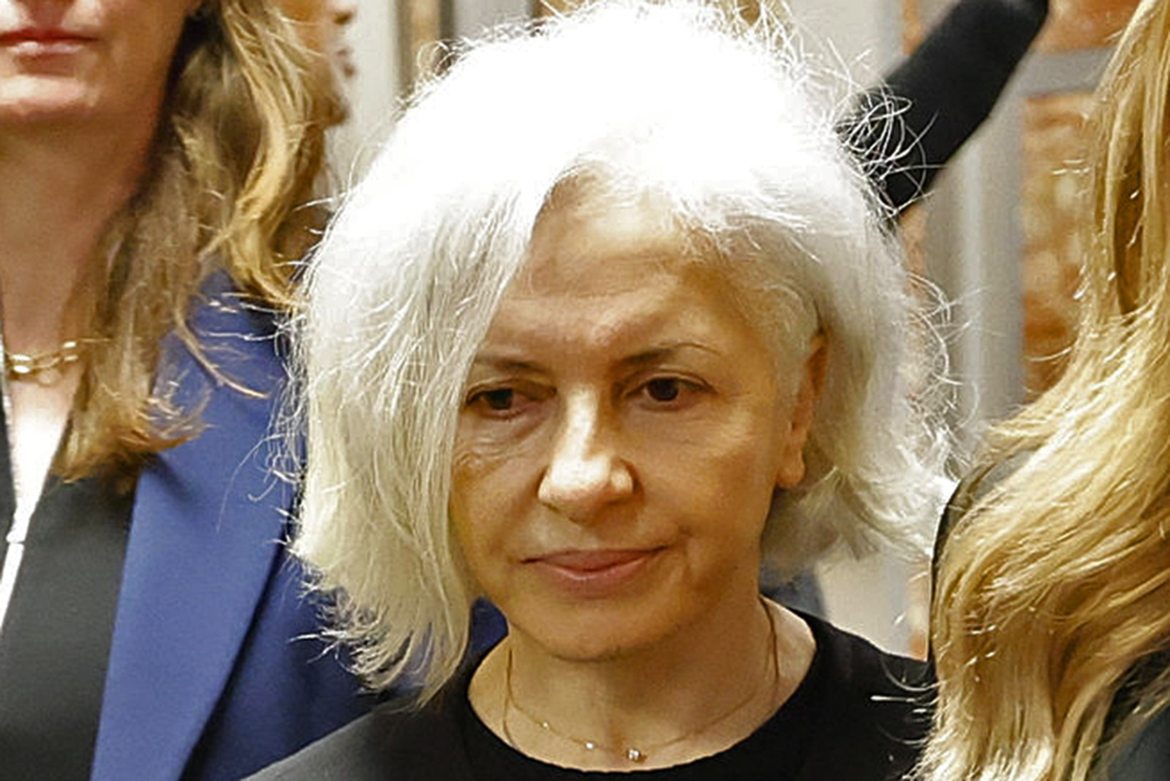 A middle-aged woman with white hair and a calm expression stands between two people with long hair, one in a blue jacket.
