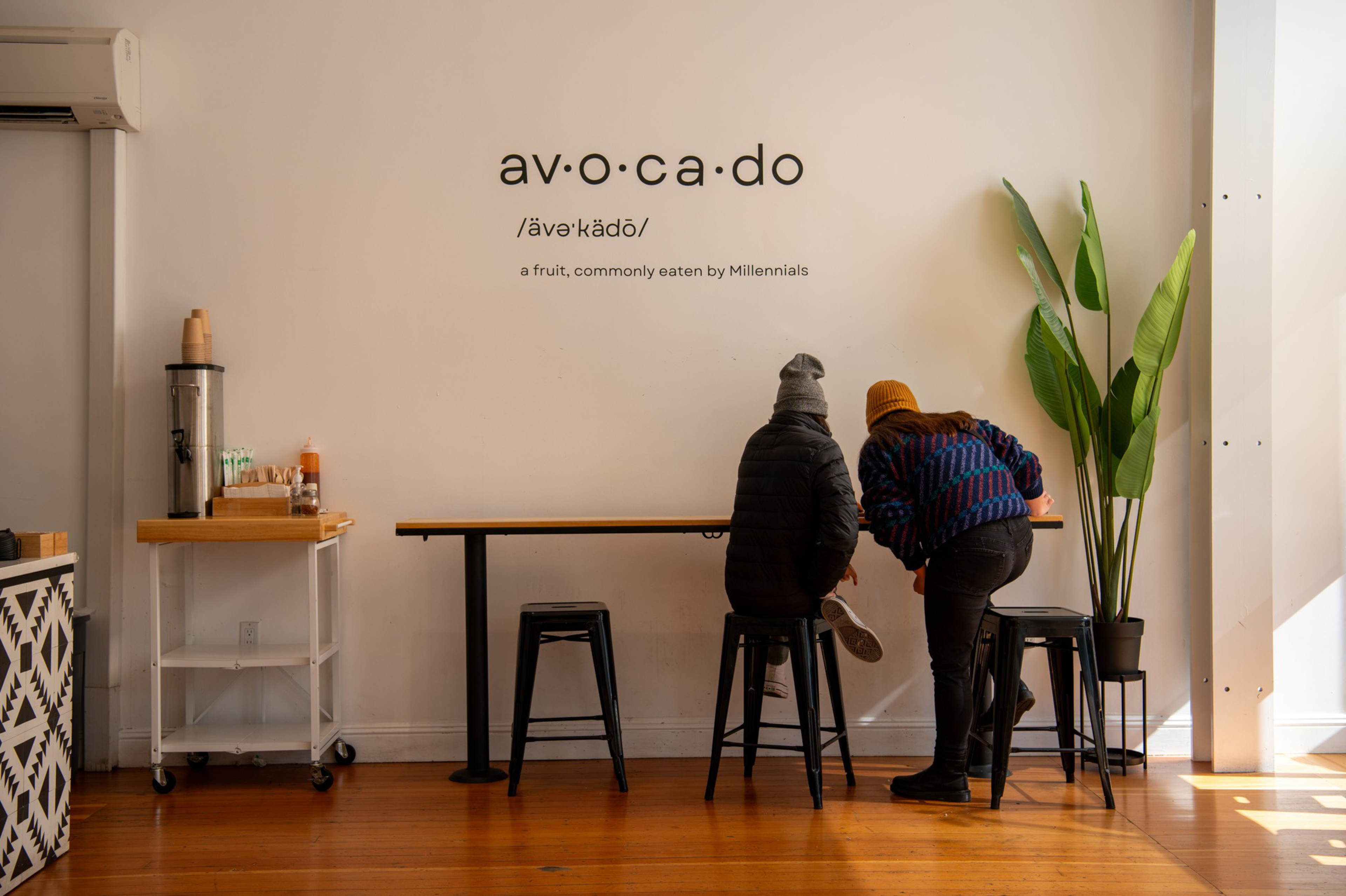 Two people sit on stools at a minimalist cafe counter with the word "avocado" and its pronunciation and definition displayed on the wall above.