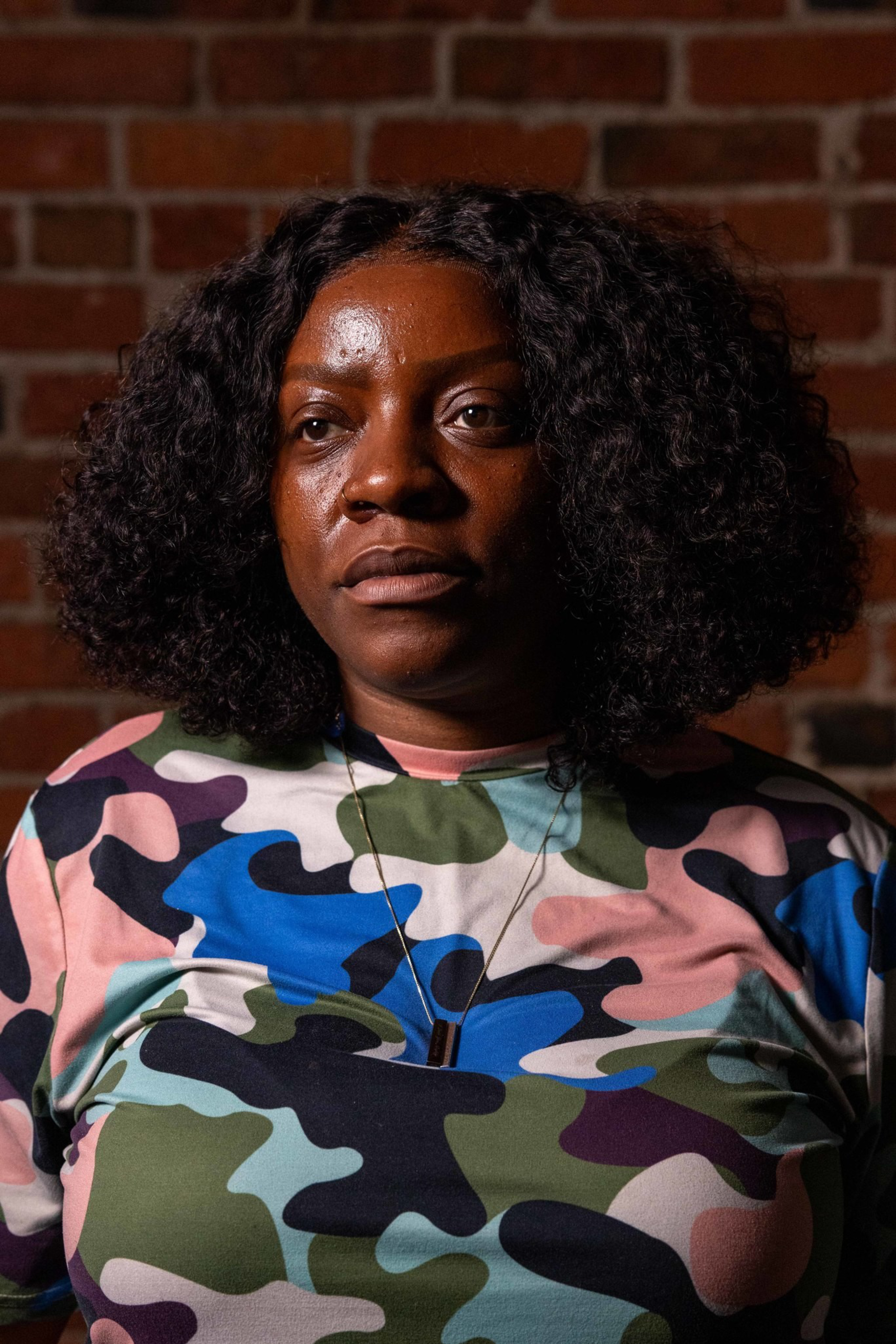 A woman with curly hair wears a colorful camouflage shirt and pendant necklace, standing against a brick wall background.