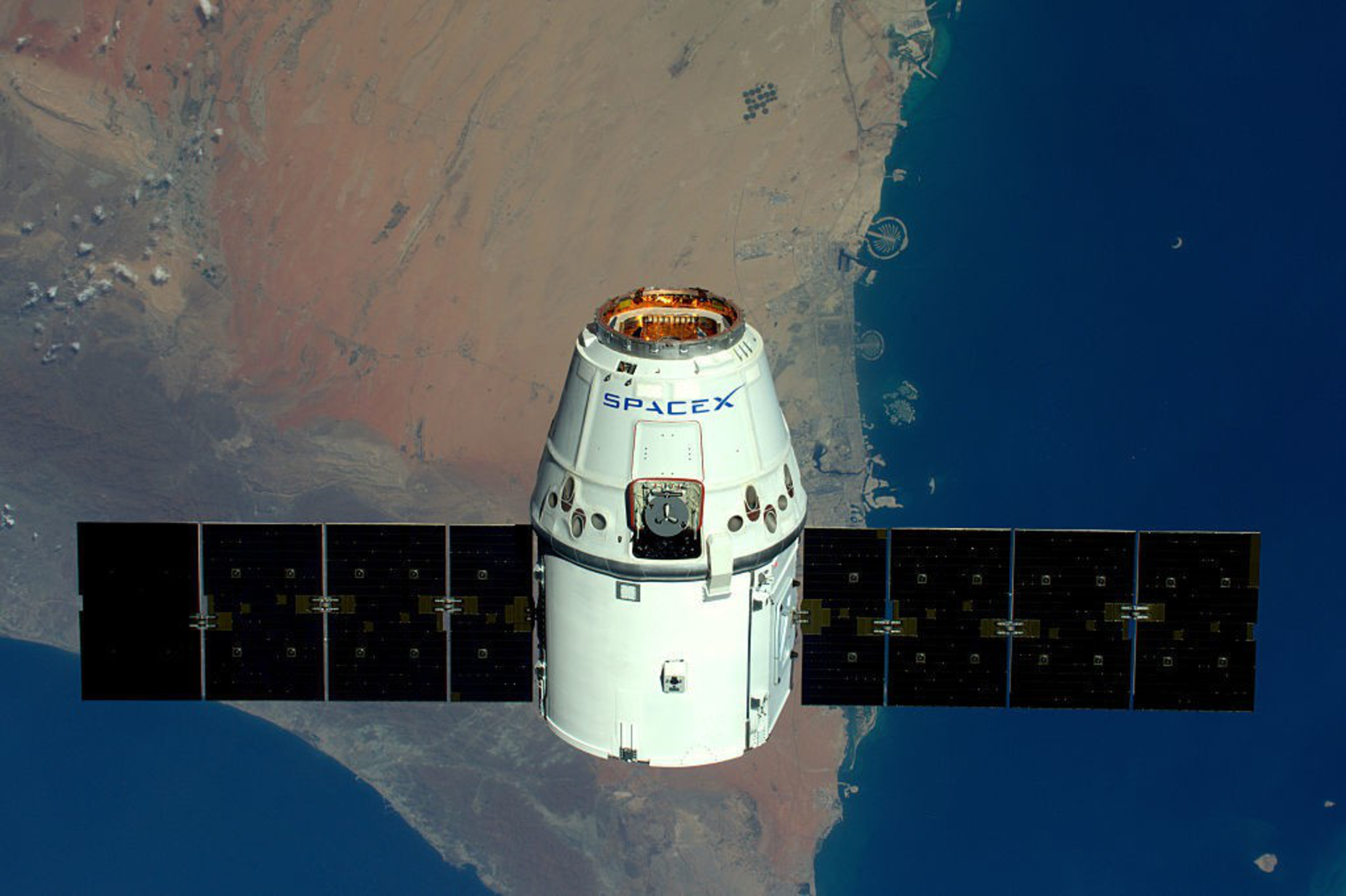 A SpaceX spacecraft orbits Earth, with vast blue waters and rugged beige land below, and a crescent moon in the distance.