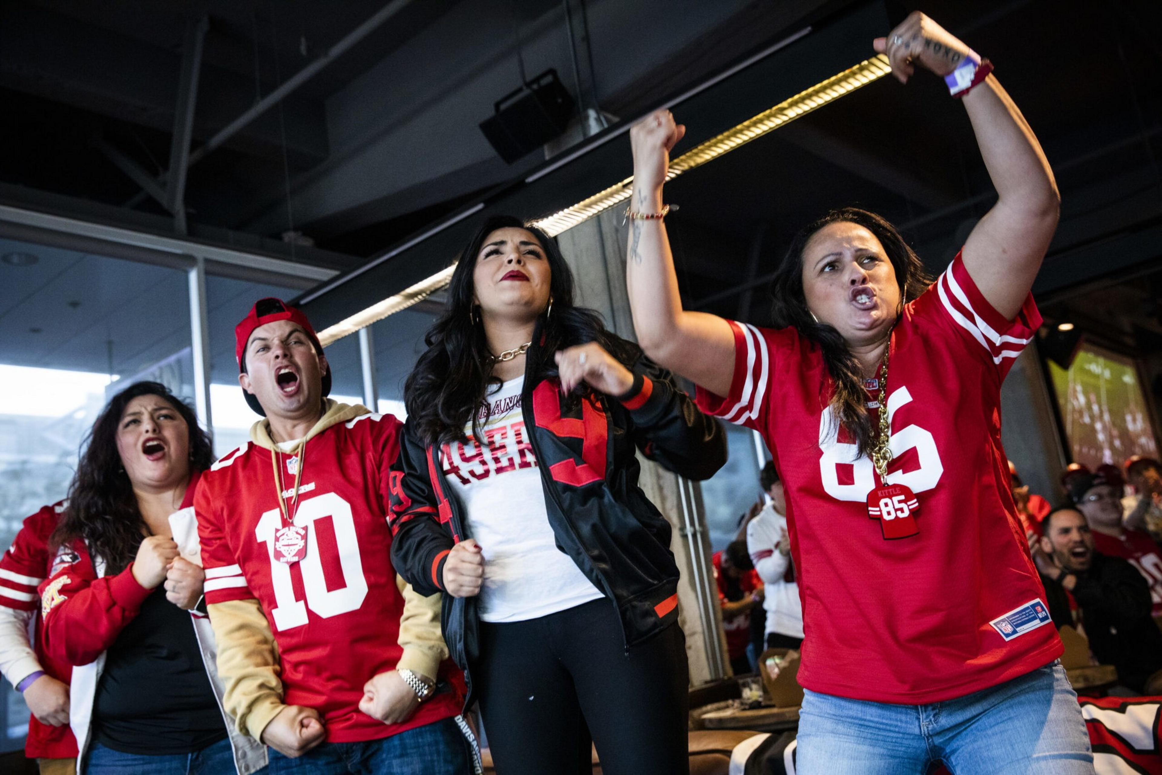 Three excited fans in red jerseys cheer at a sporting event, with one holding a rope.