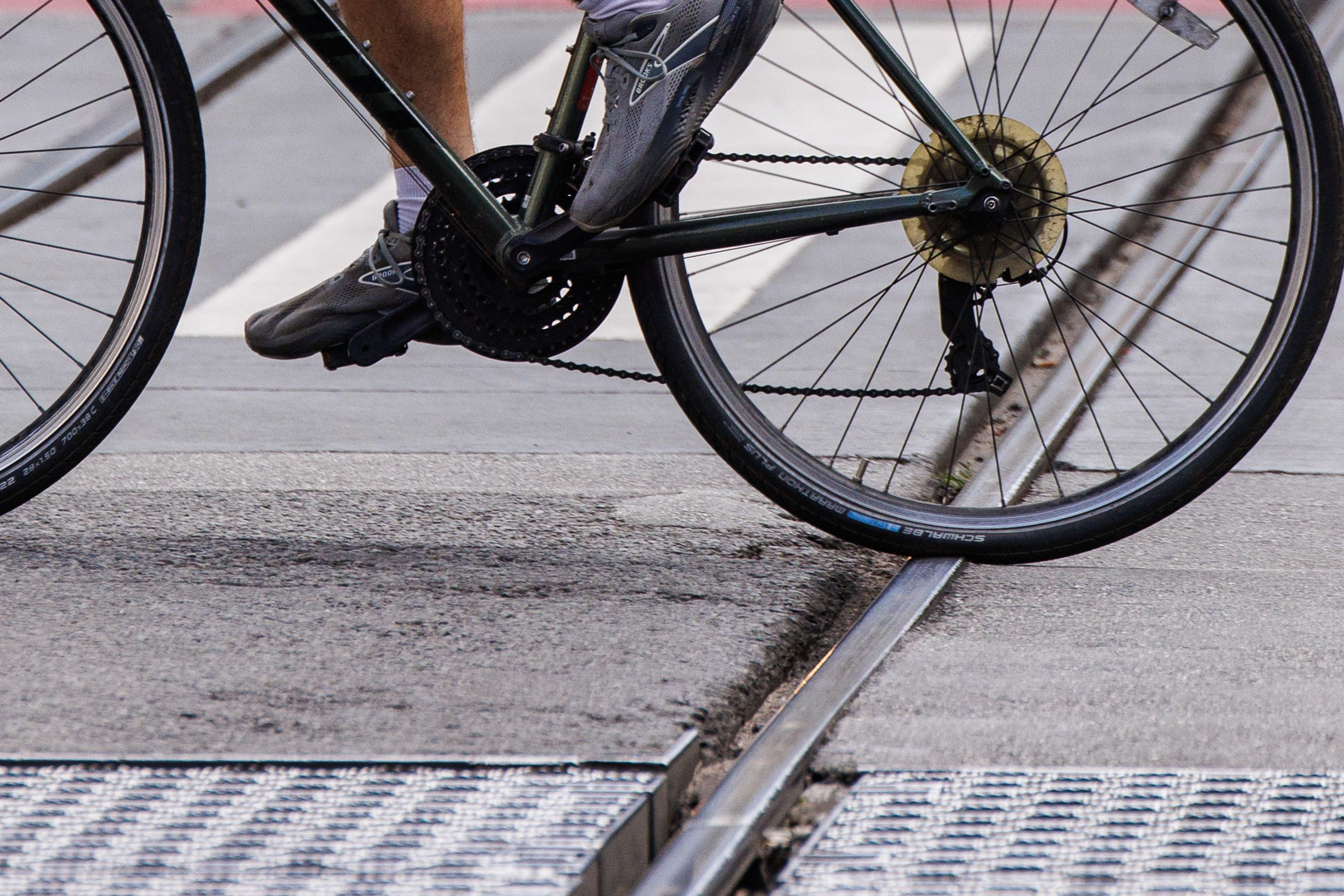 A person wearing gray sneakers pedals a bicycle crossing over a metal rail embedded in the pavement. The bike tire is directly above the rail.
