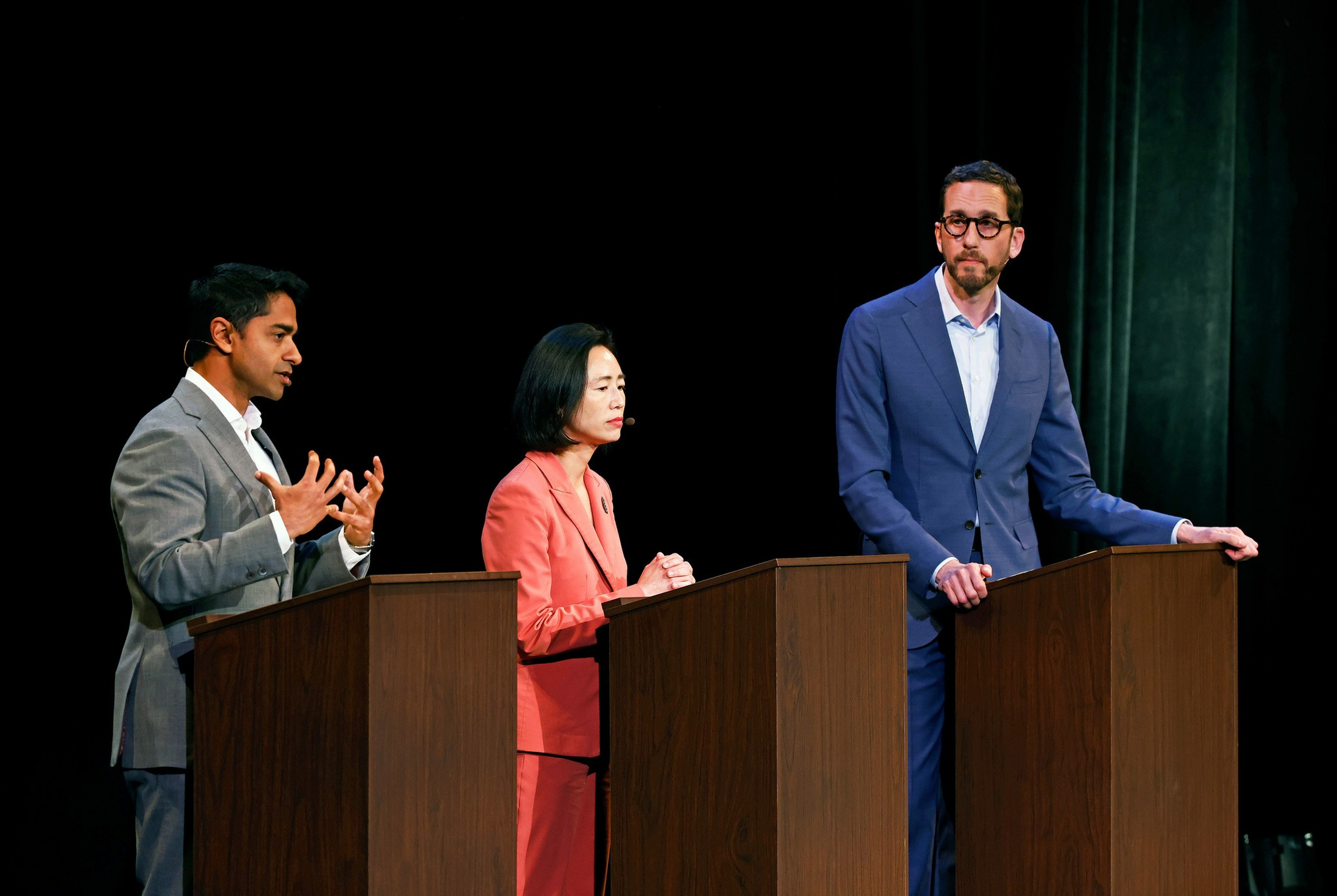 Three people stand behind wooden podiums on a stage, with one man speaking and gesturing while the others listen attentively.