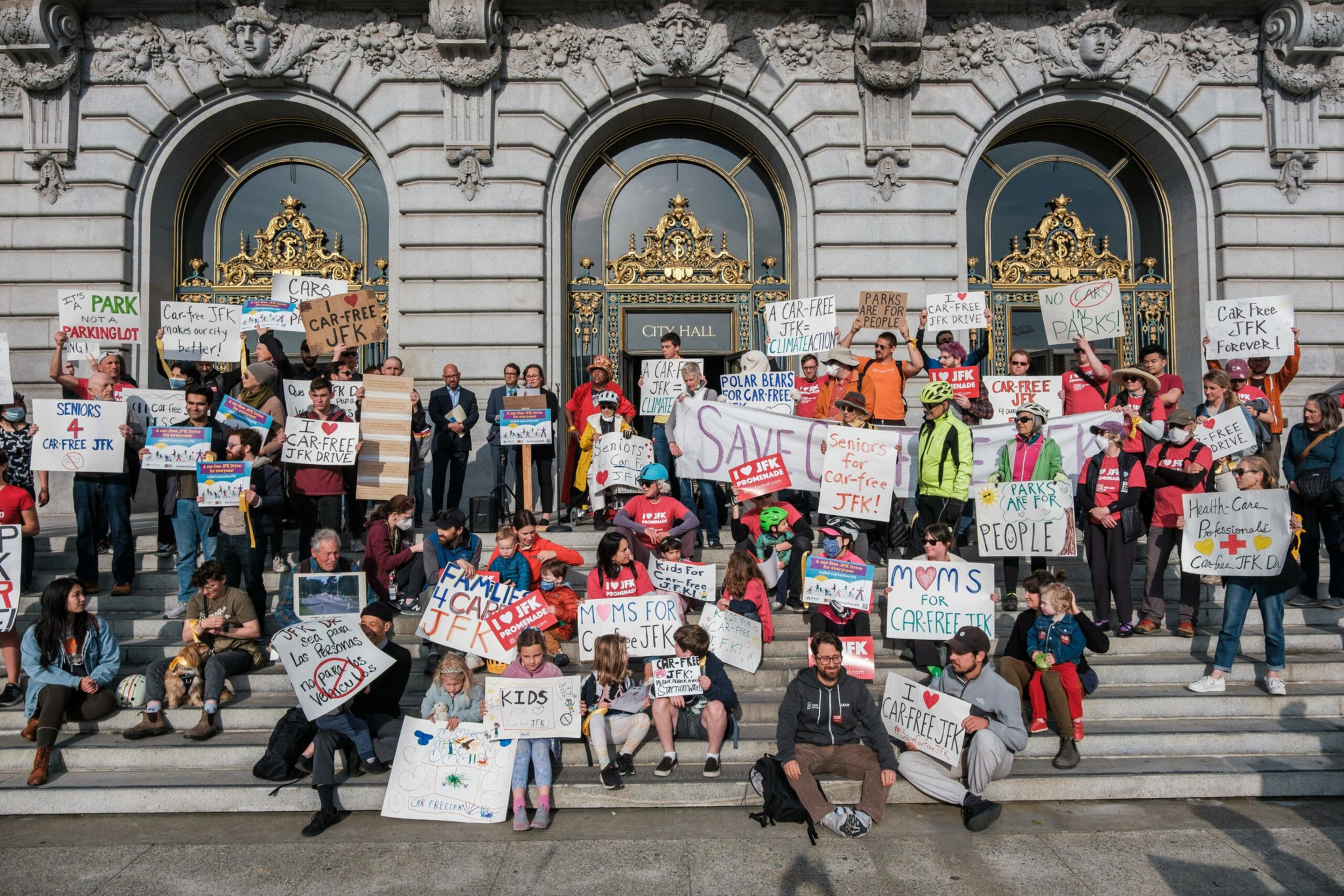 A diverse group of people on ornate city hall steps hold signs advocating for a car-free JFK Drive, emphasizing parks, families, and climate action.