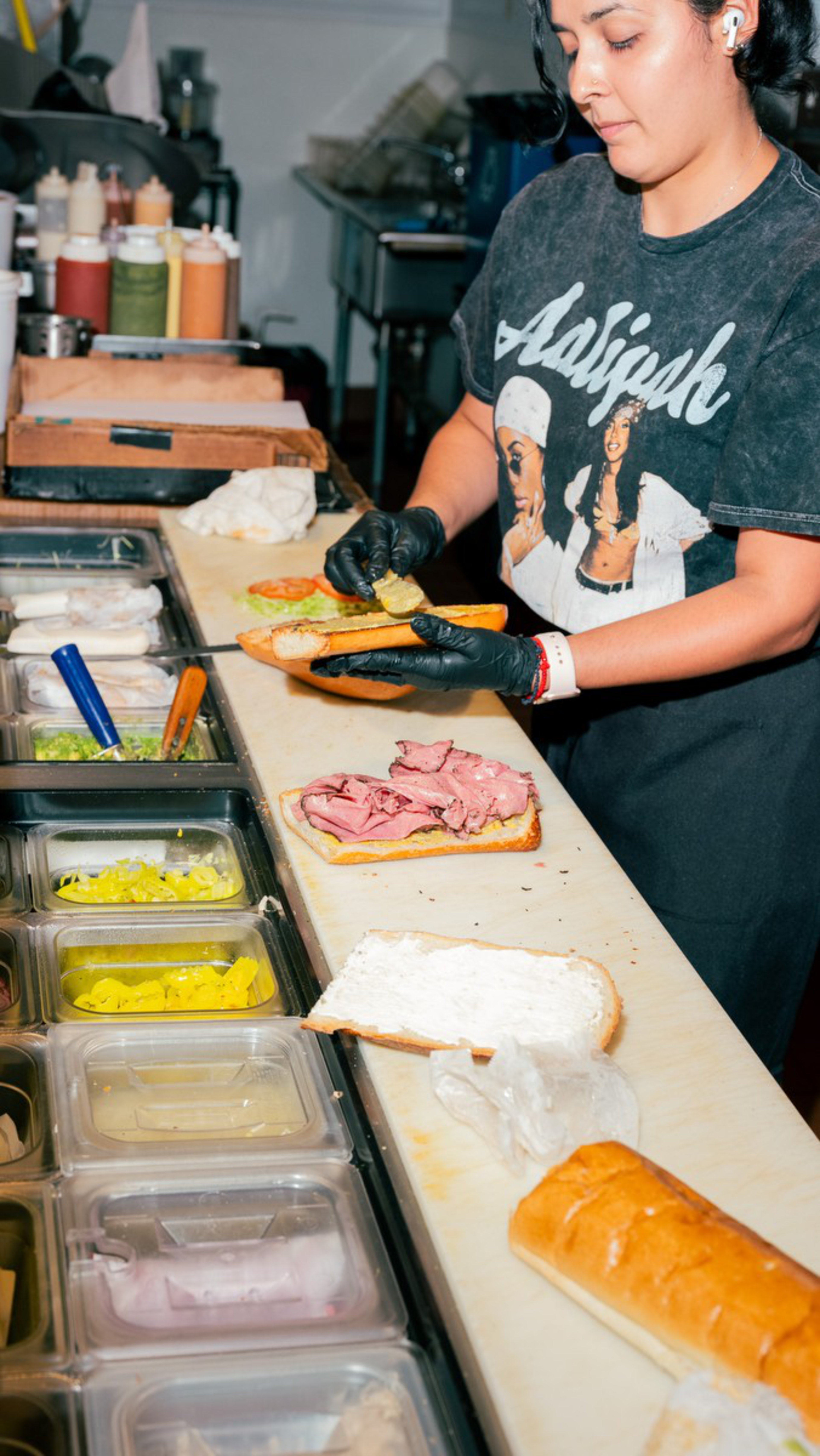 A person wearing black gloves prepares sandwiches at a deli counter with various sandwich ingredients and sauces in containers.