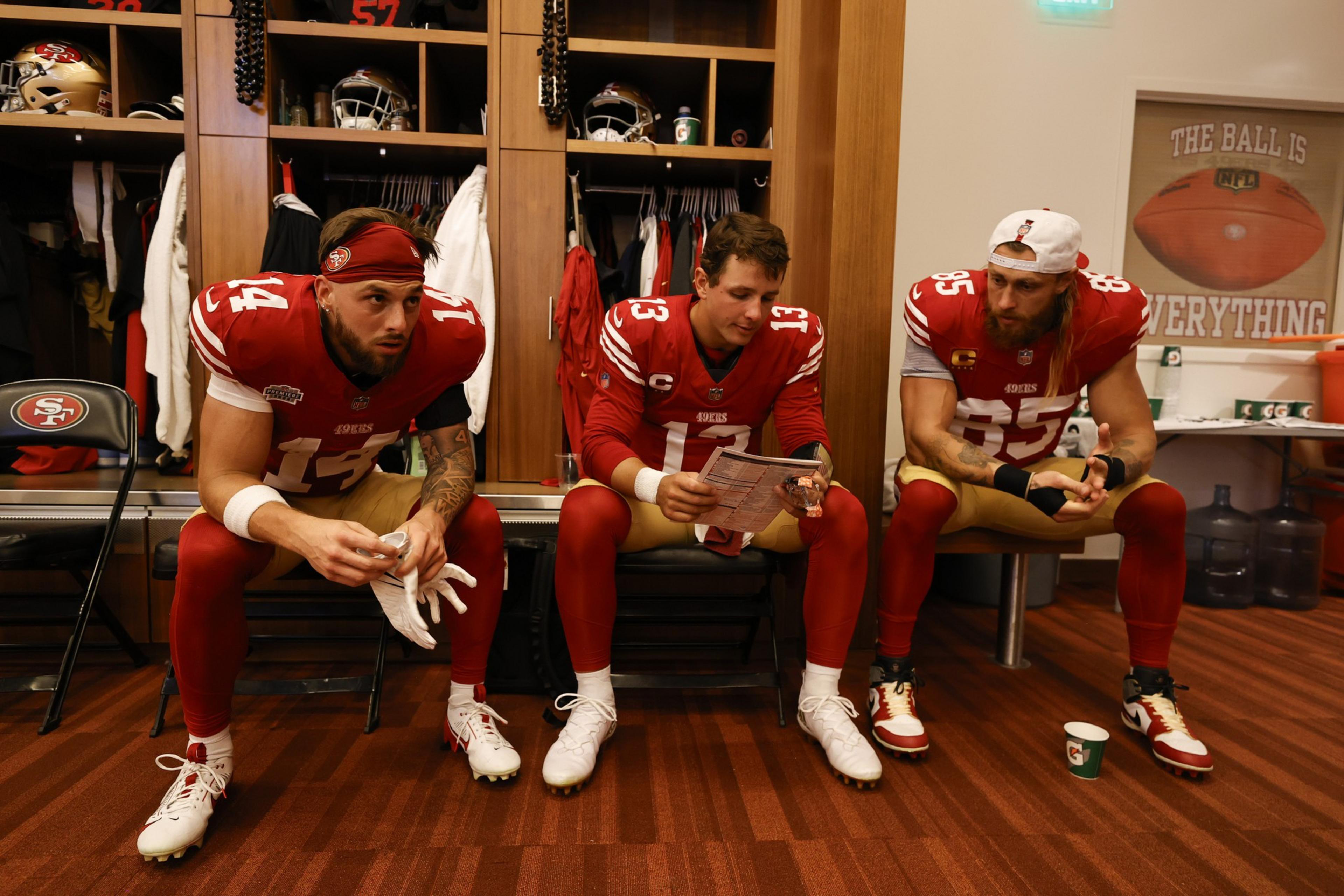 Three football players in red uniforms sit in a locker room, two hold papers, and one adjusts his gloves. A motivational poster is on the wall.