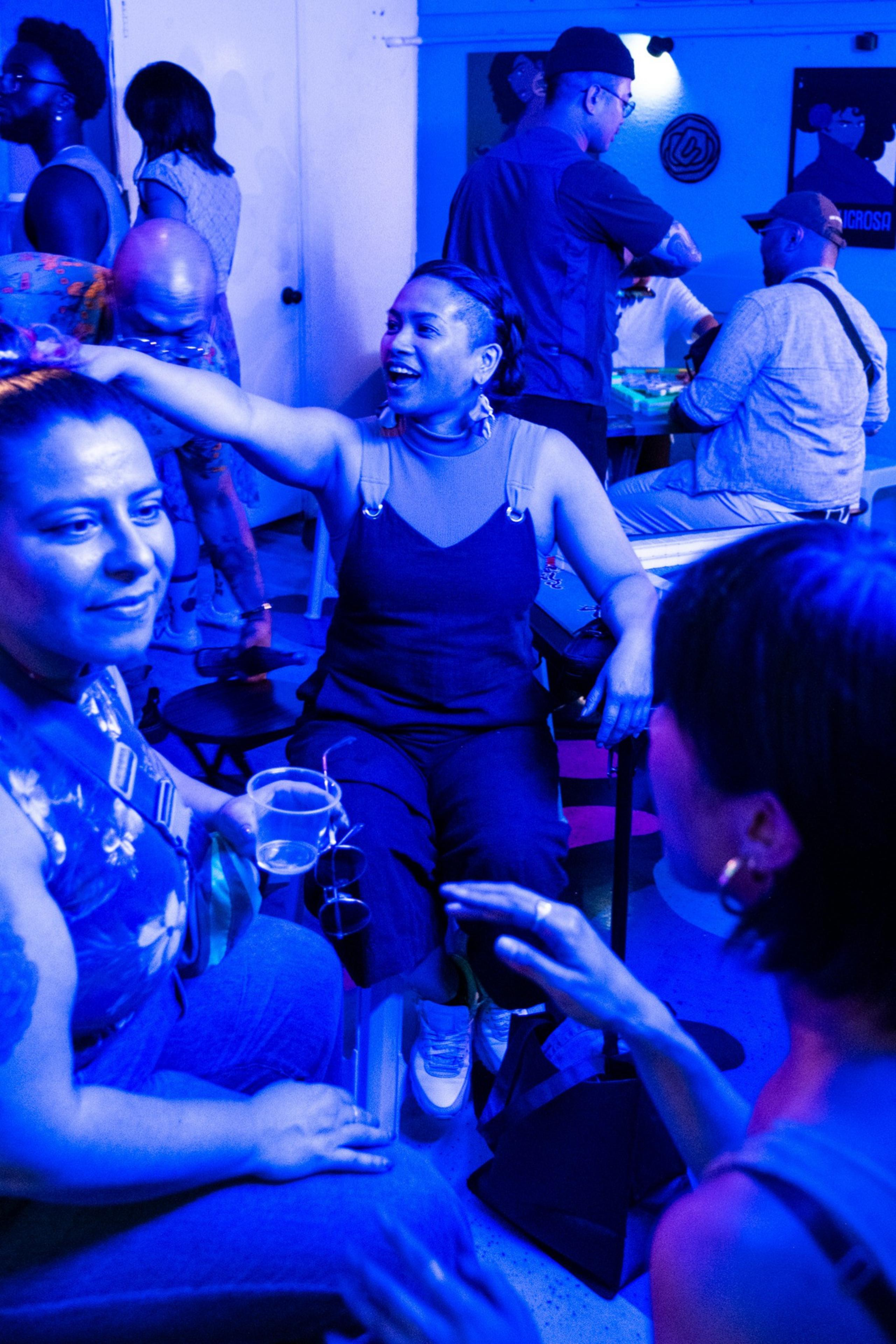 People are in a dimly lit room with blue lighting, engaging in conversation. One person is drinking from a cup, and others are interacting energetically.