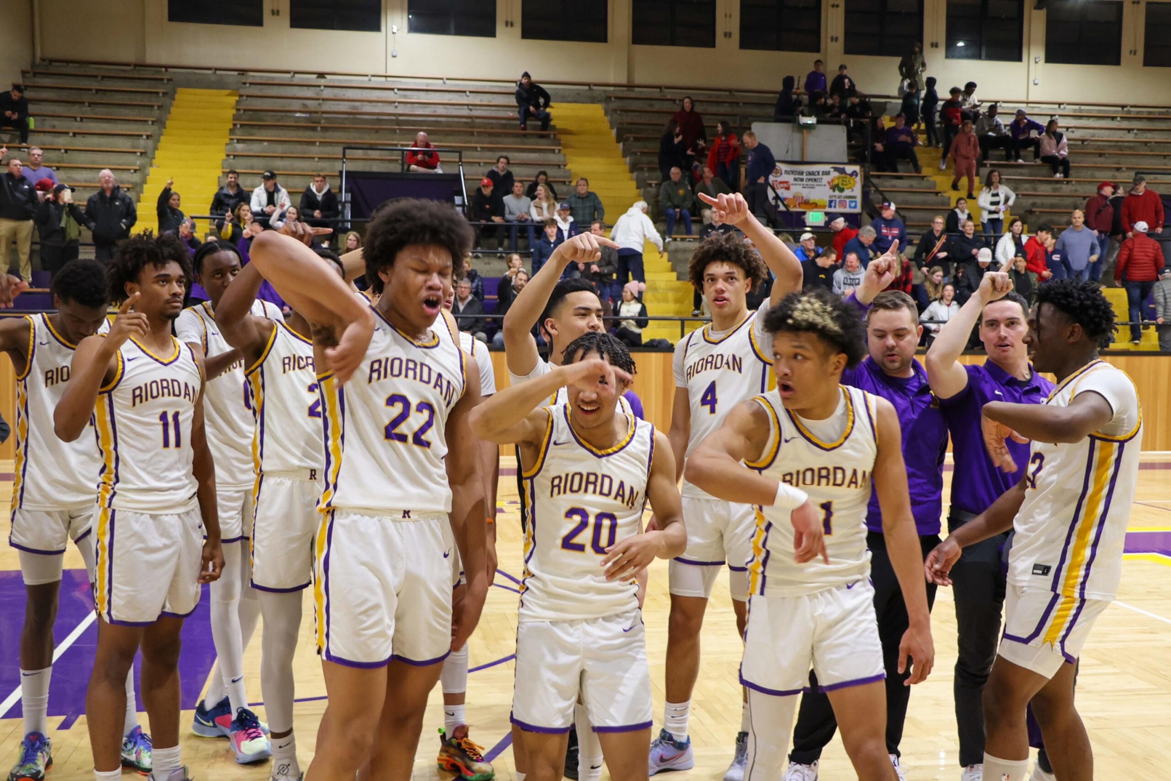 A basketball team in white "Riordan" uniforms is celebrating on the court with some players raising their arms and cheering.