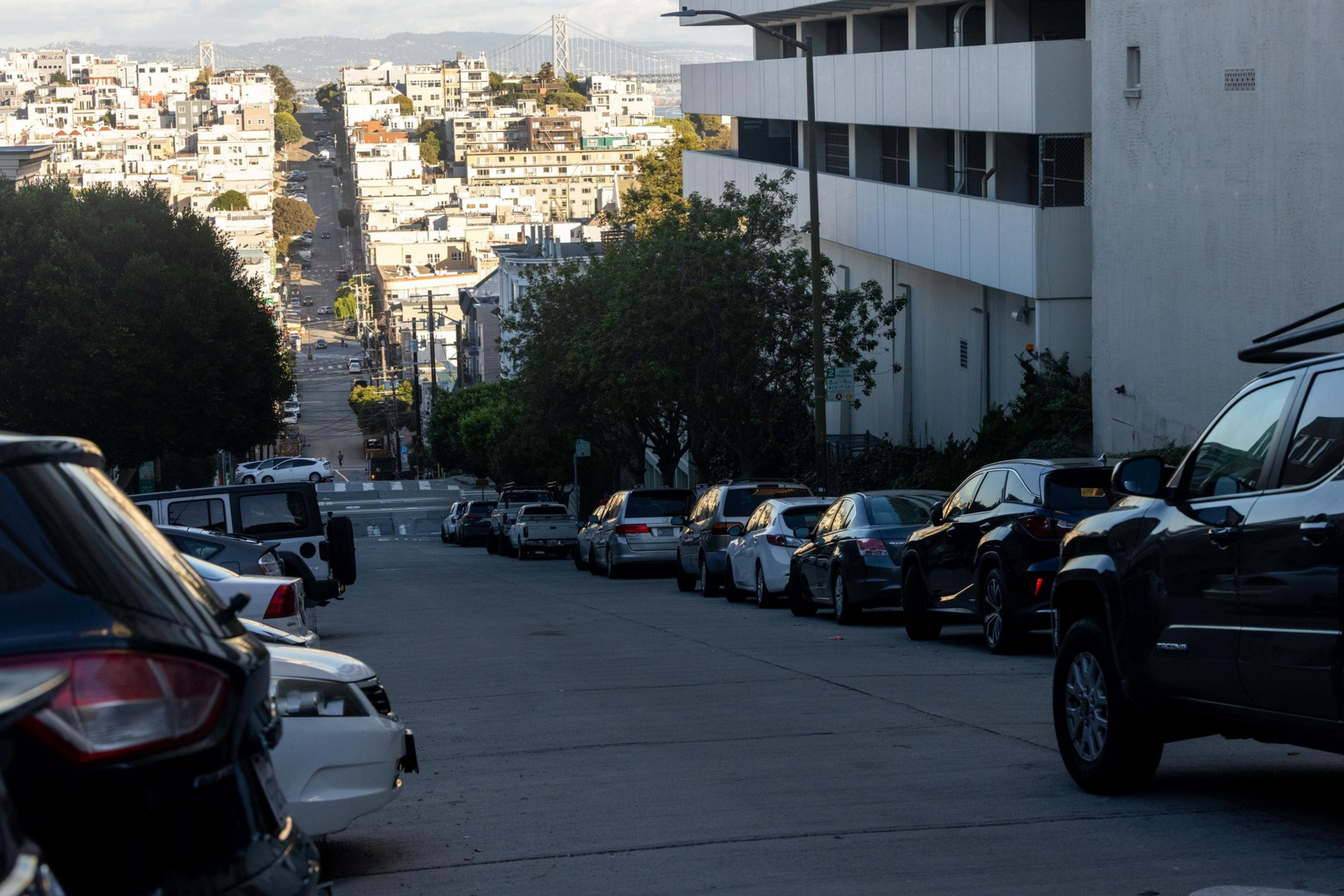 A steep urban street lined with parked cars on both sides, trees, and buildings, with a bridge visible in the distant background.