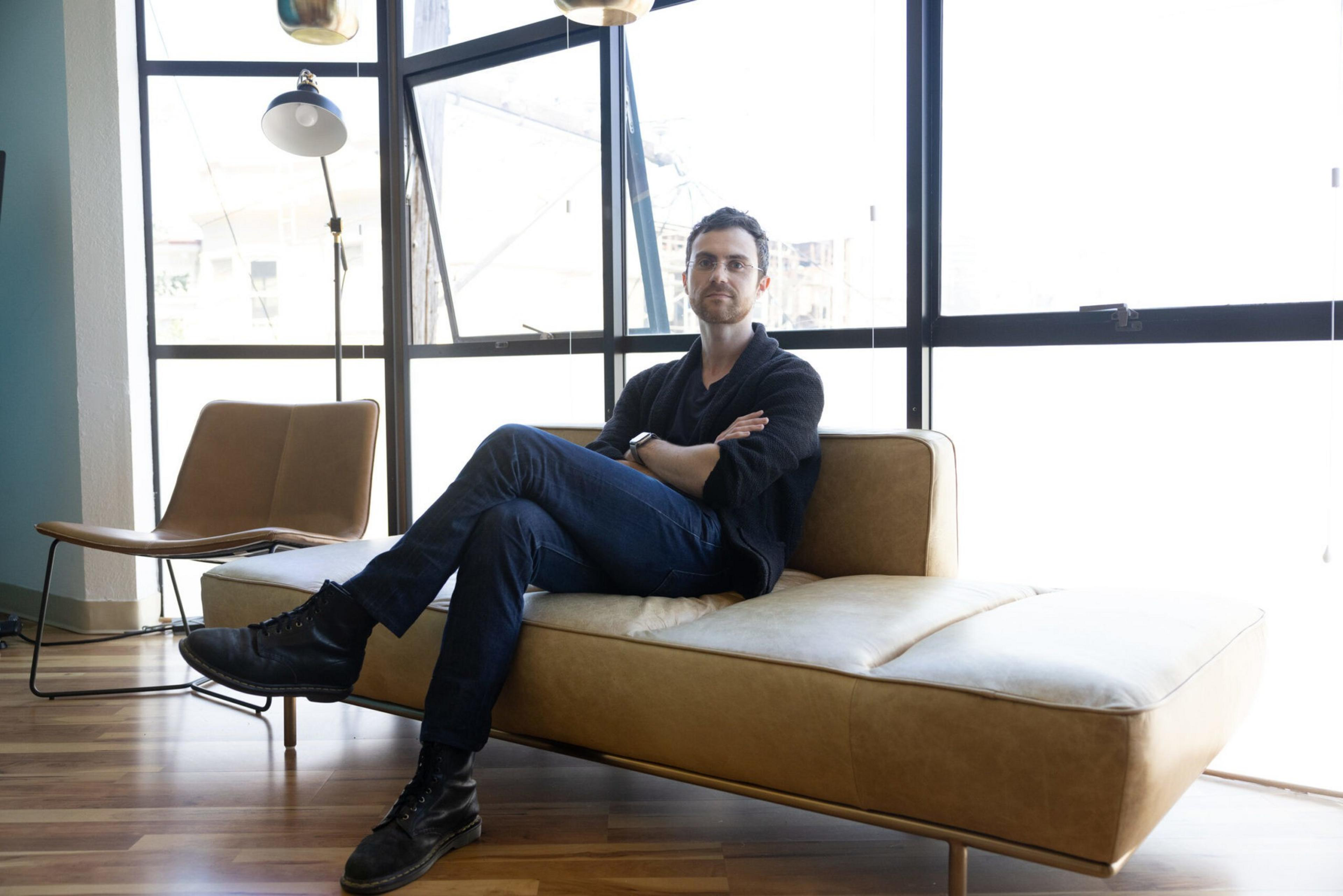 A man with glasses, dark hair, and a beard sits cross-legged on a tan leather sofa, wearing dark jeans and a black shirt by large windows.