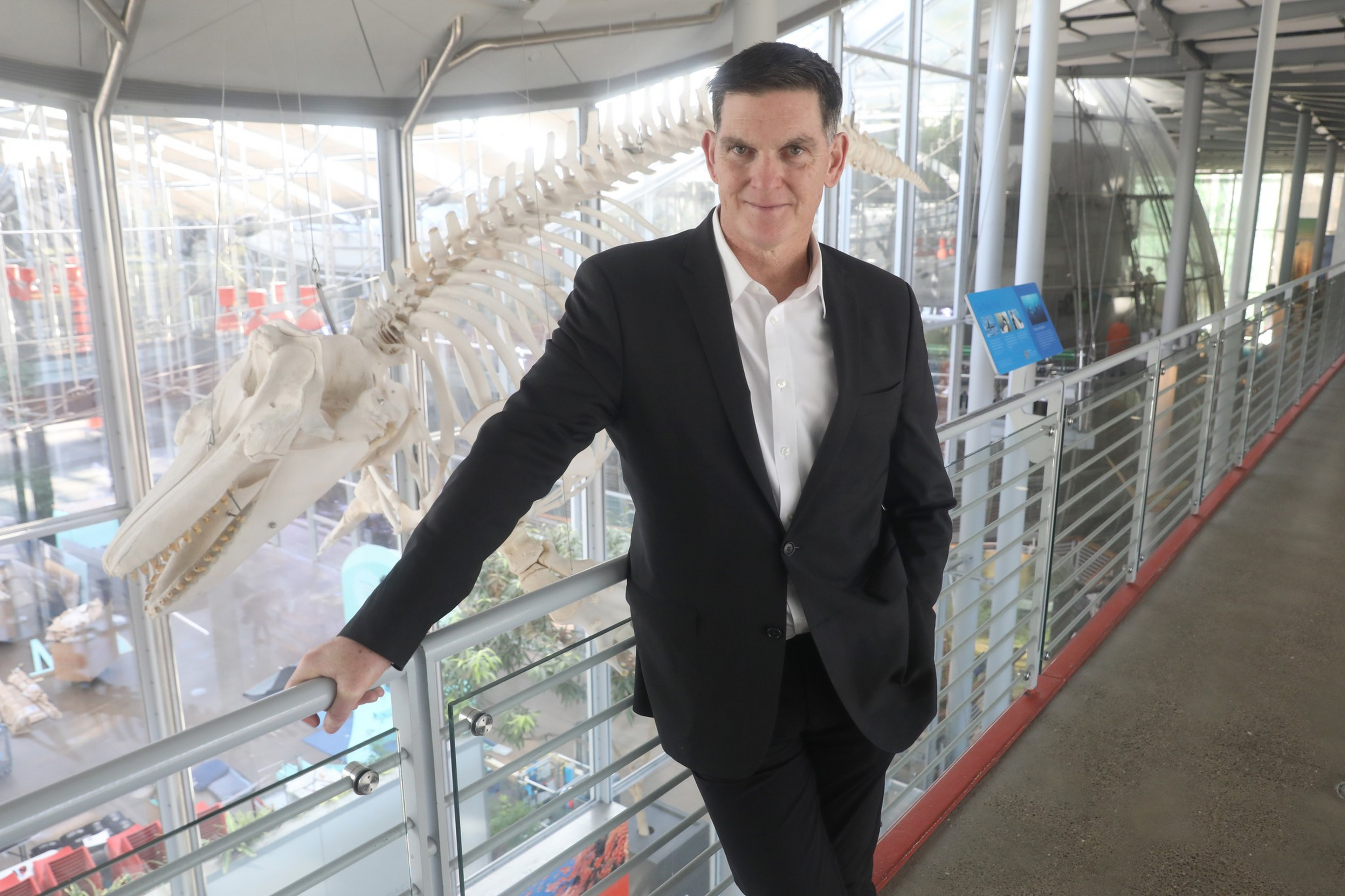 A man in a black suit stands with one hand on a railing inside a modern building, with a large whale skeleton suspended behind him.
