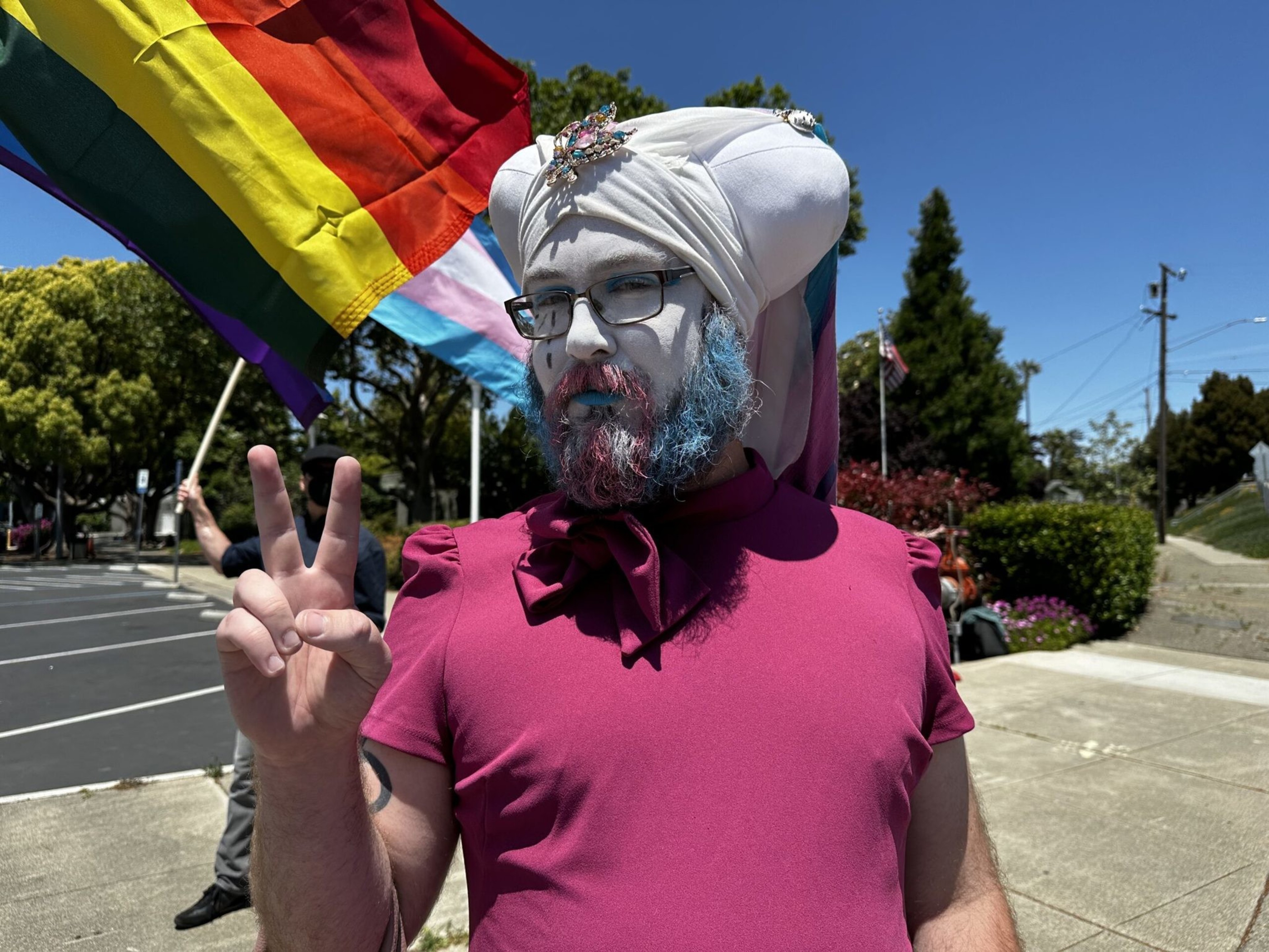 A person with face paint and colorful beard wears a white turban and magenta top, holding up a peace sign, with pride flags in the background.