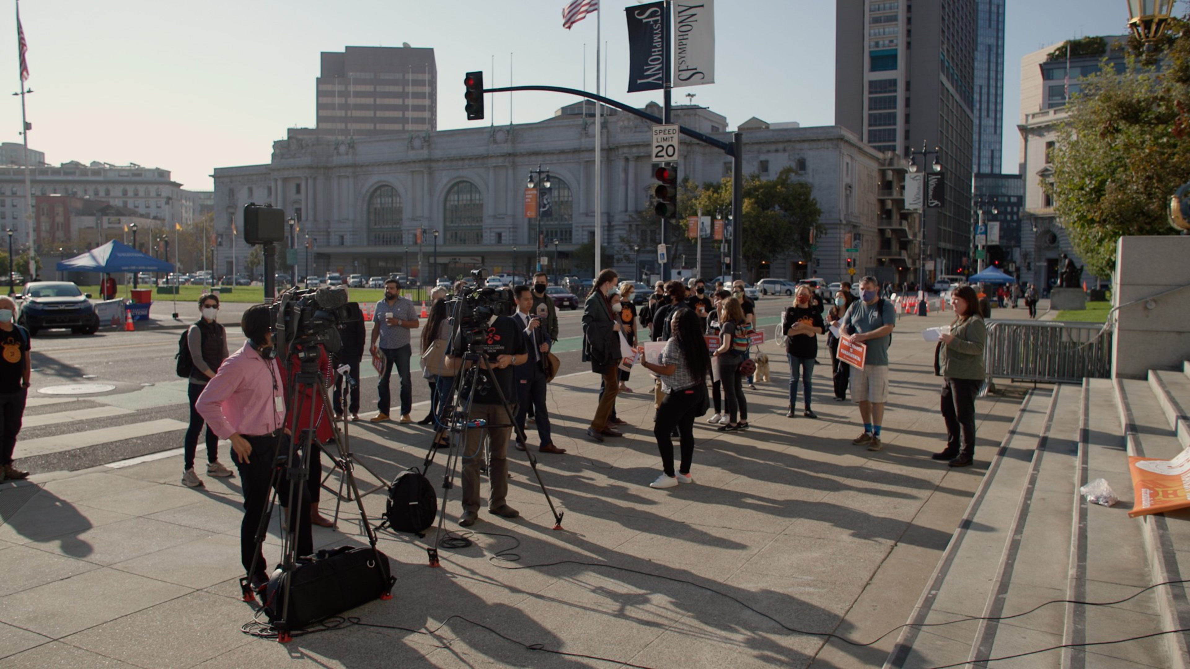 A small group of people, some with cameras and tripods, gather near city steps on a sunny day, with buildings and traffic lights in the background.