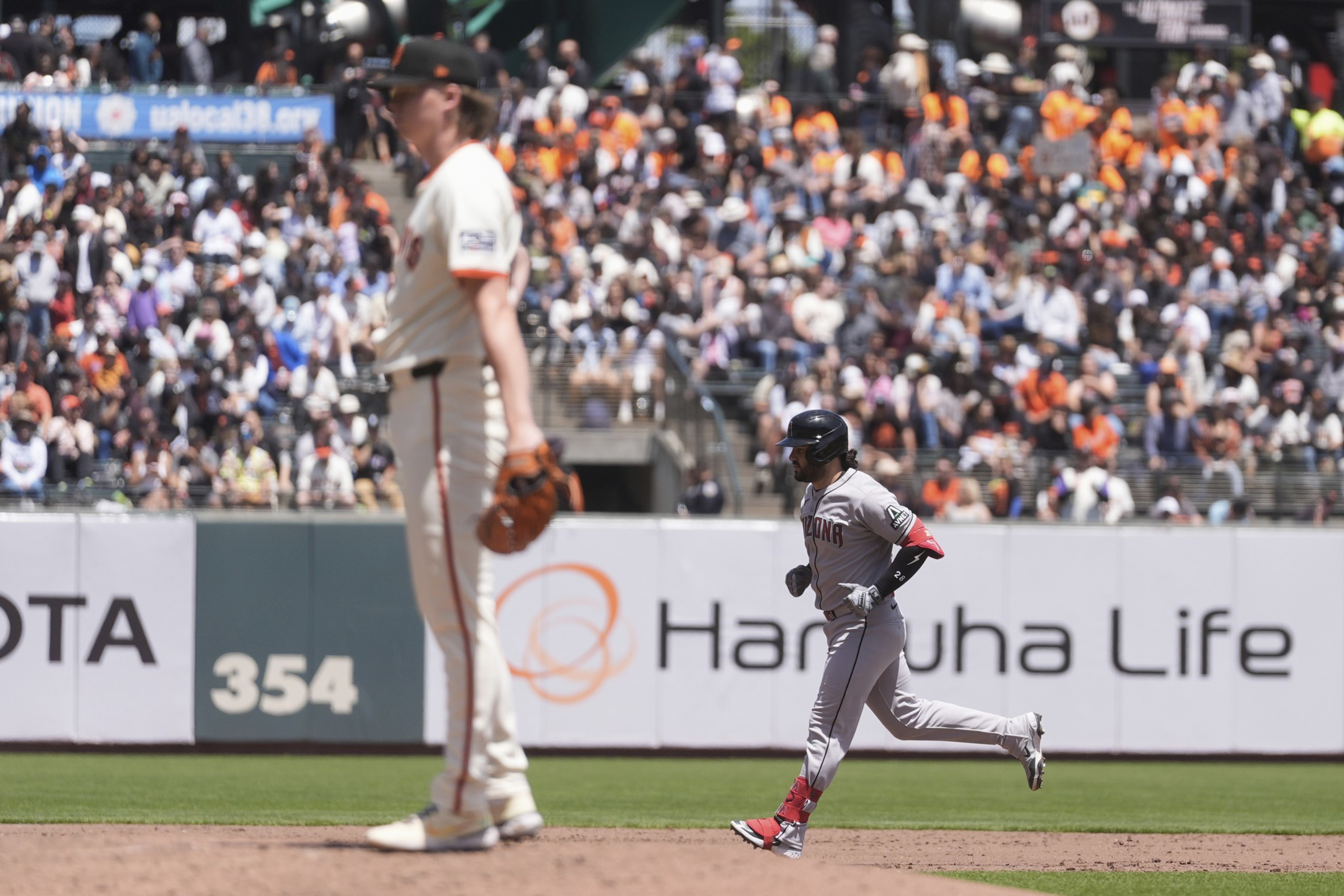 A baseball player in a gray uniform runs past a pitcher in a white uniform with an orange cap. The stands are filled with fans, some wearing orange shirts.