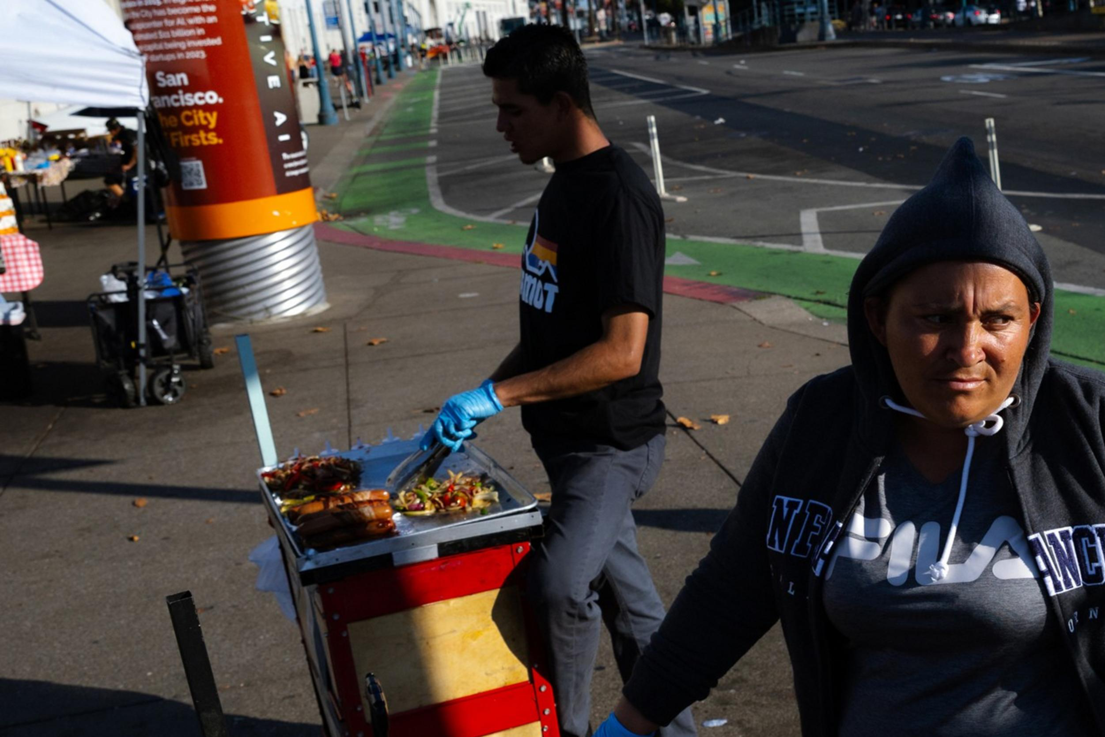 A woman in a hoodie stands next to a food cart with grilled sausages and onions. Another person is preparing food in the background on a city street.