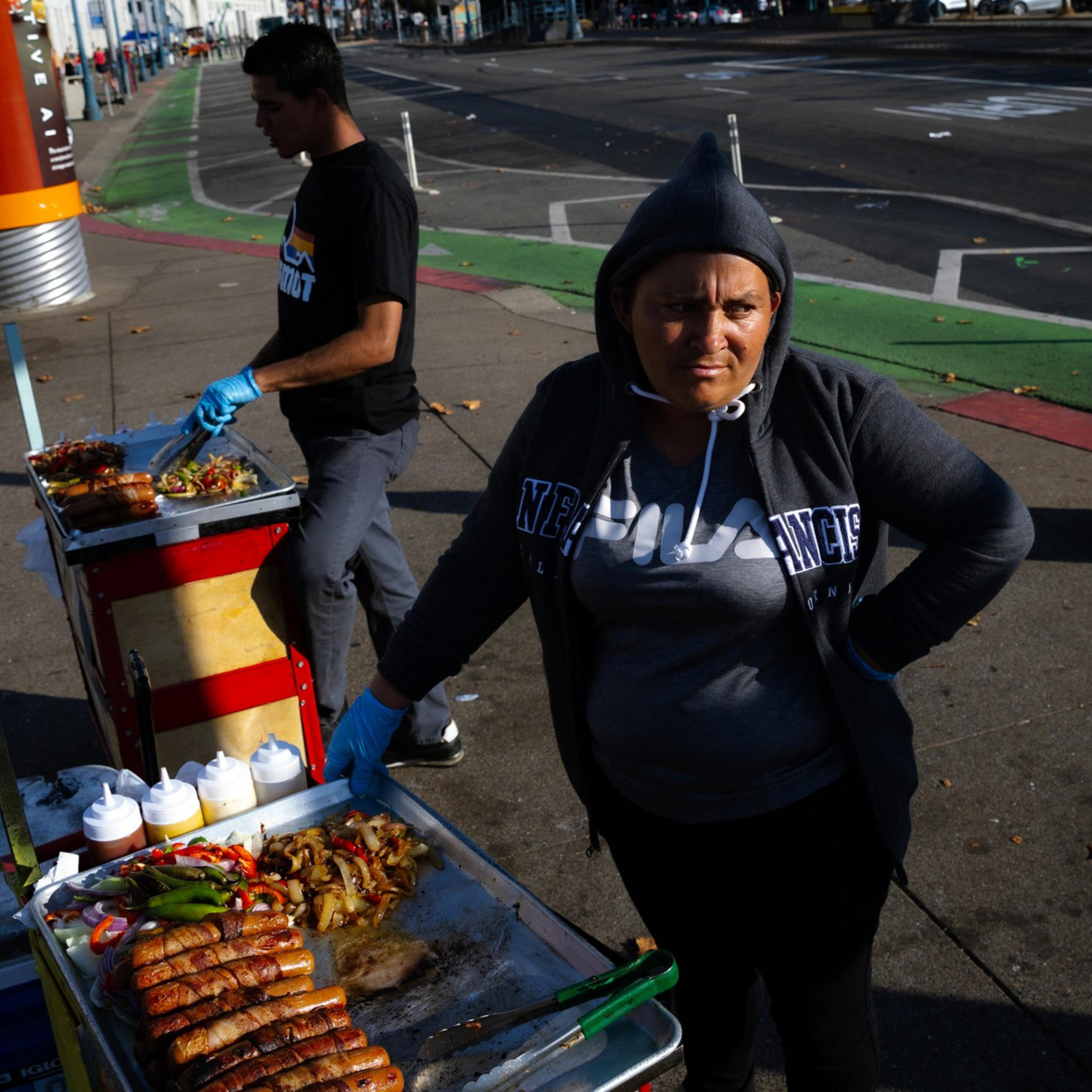 A woman in a hoodie stands next to a food cart with grilled sausages and onions. Another person is preparing food in the background on a city street.
