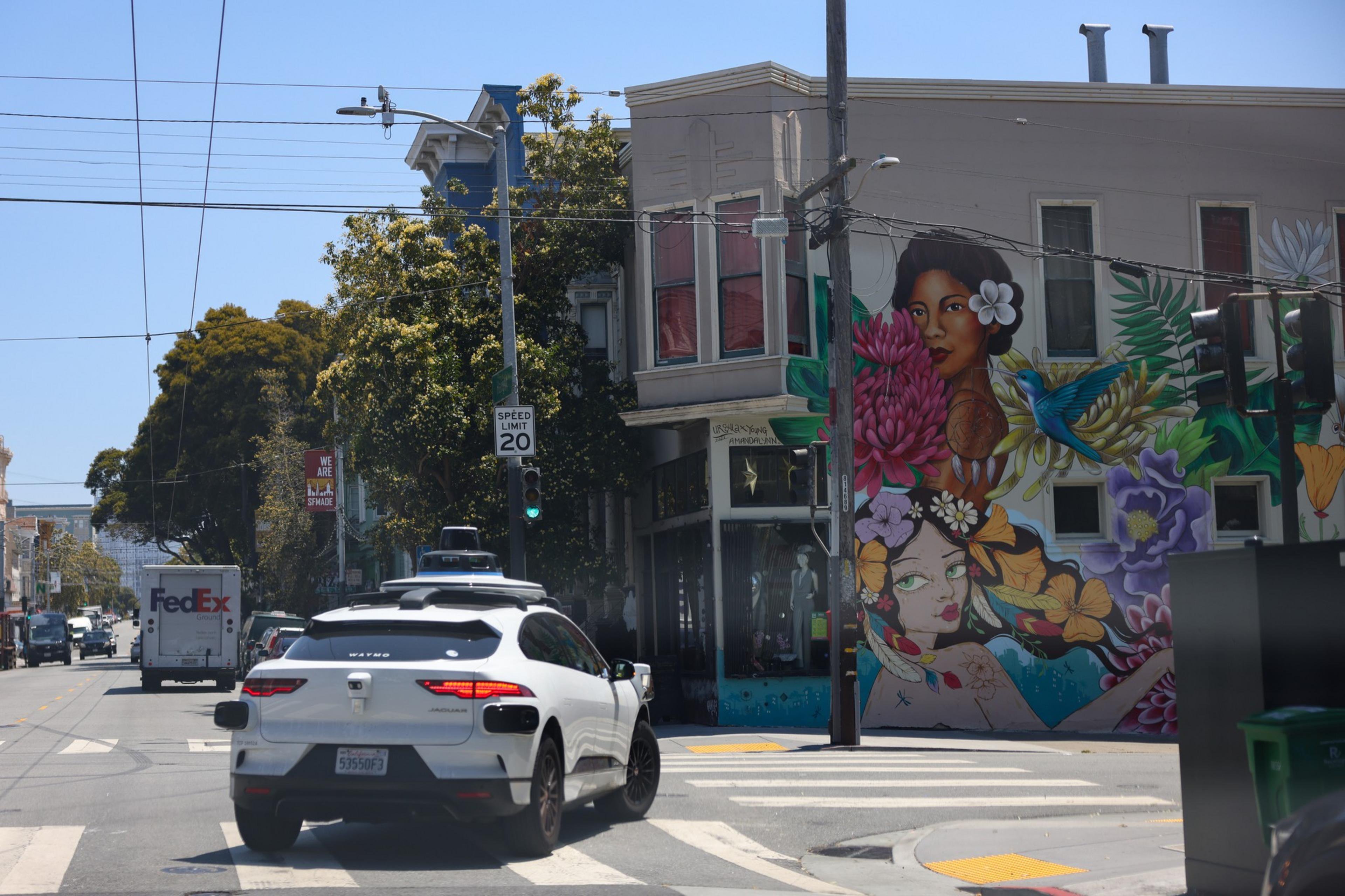 A white car waits at a traffic light near a colorful mural of two women with flowers and a hummingbird on a building corner.