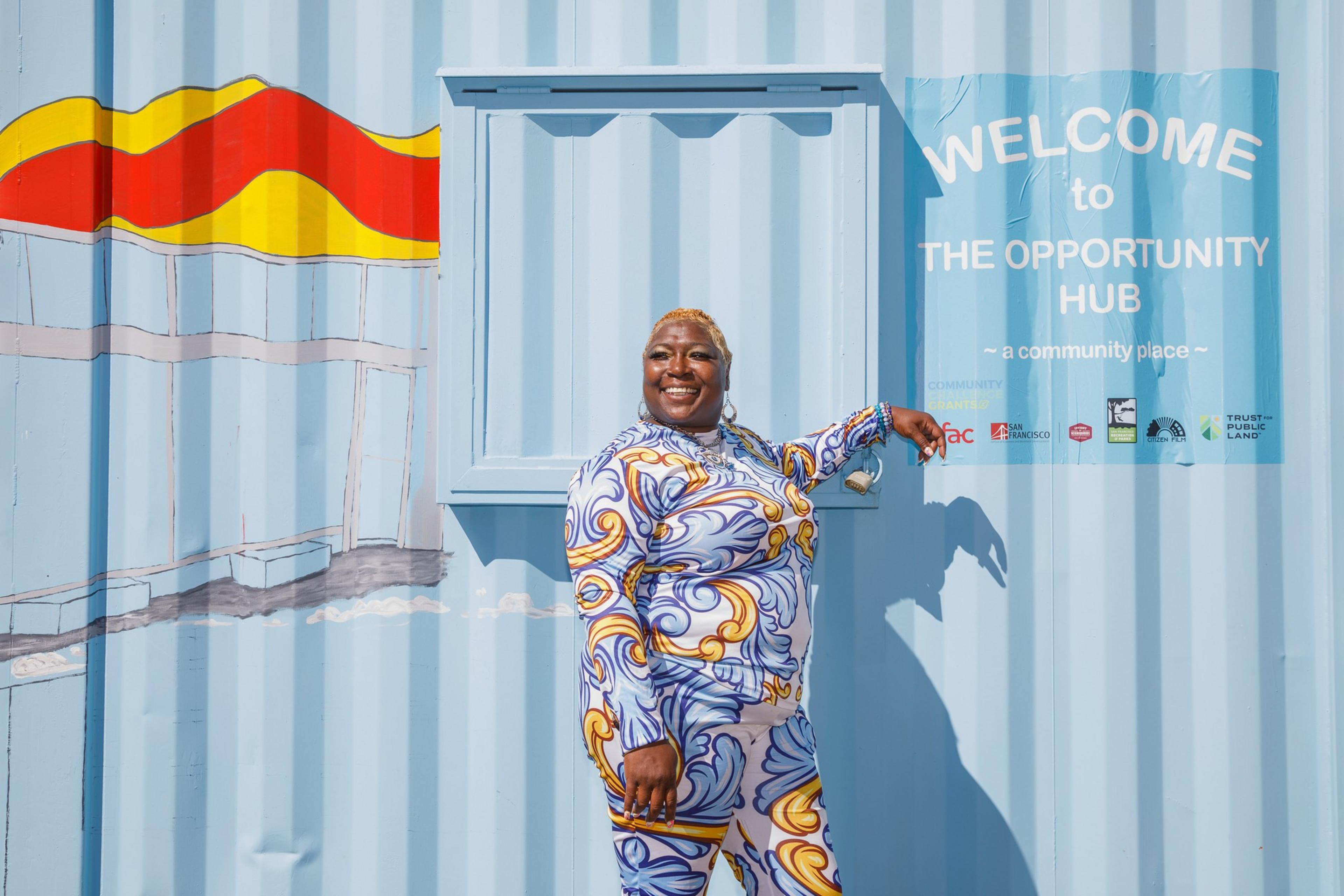 A smiling person in a bold print outfit stands by a blue container labeled &quot;Opportunity Hub.&quot;