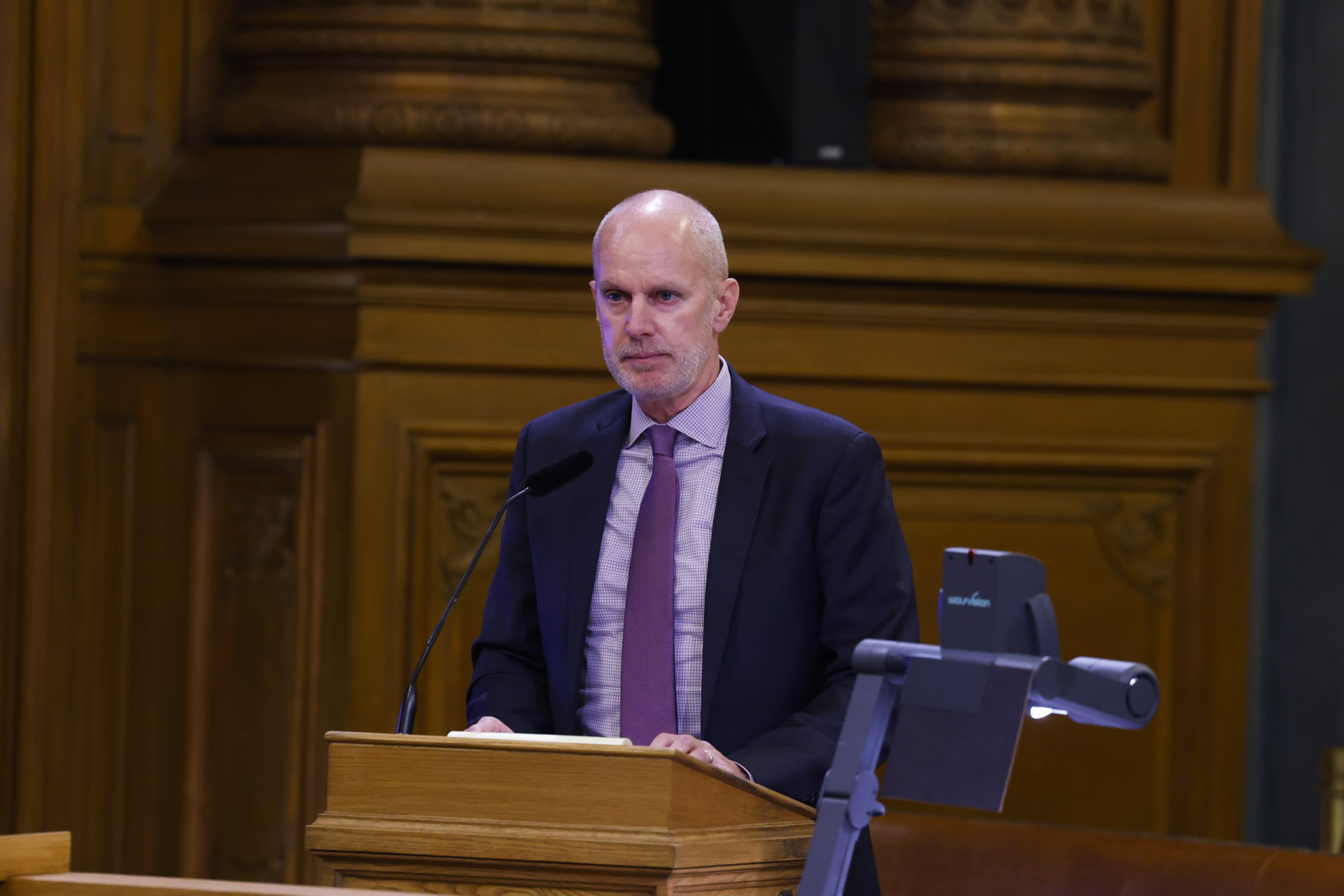 A man in a suit and tie stands at a wooden podium, speaking into a microphone, in a formal setting with ornate wood paneling in the background.