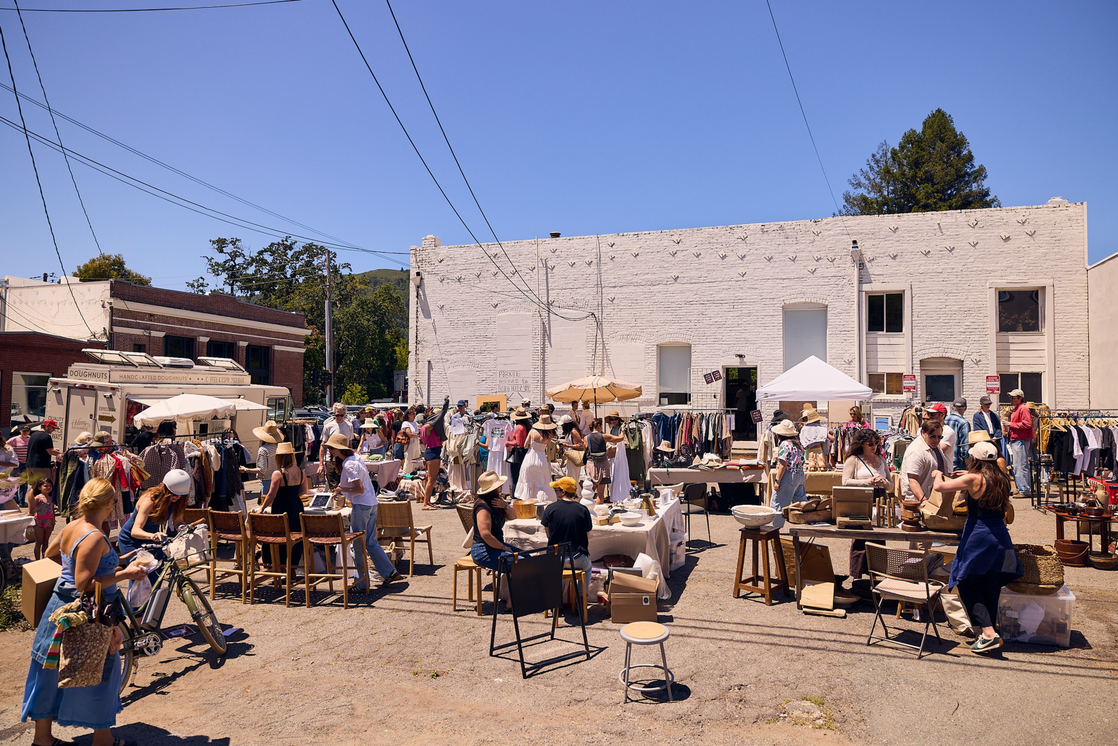 A busy outdoor market with people browsing clothing racks, tables with items for sale, and a food truck under a clear blue sky.
