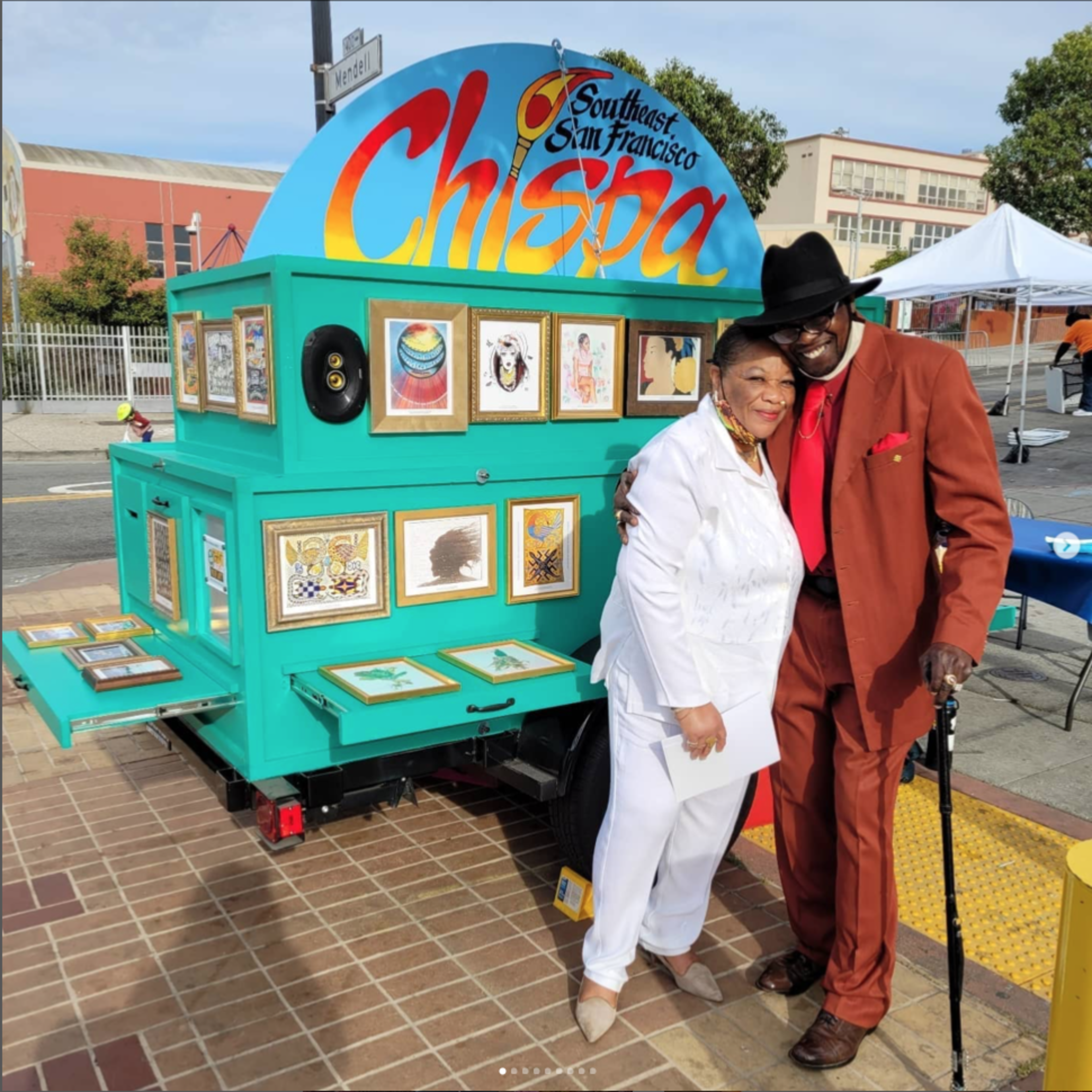 A man in a rust-colored suit and black hat hugs a woman in a white outfit in front of a colorful art display labeled "Chispa Southeast San Francisco."
