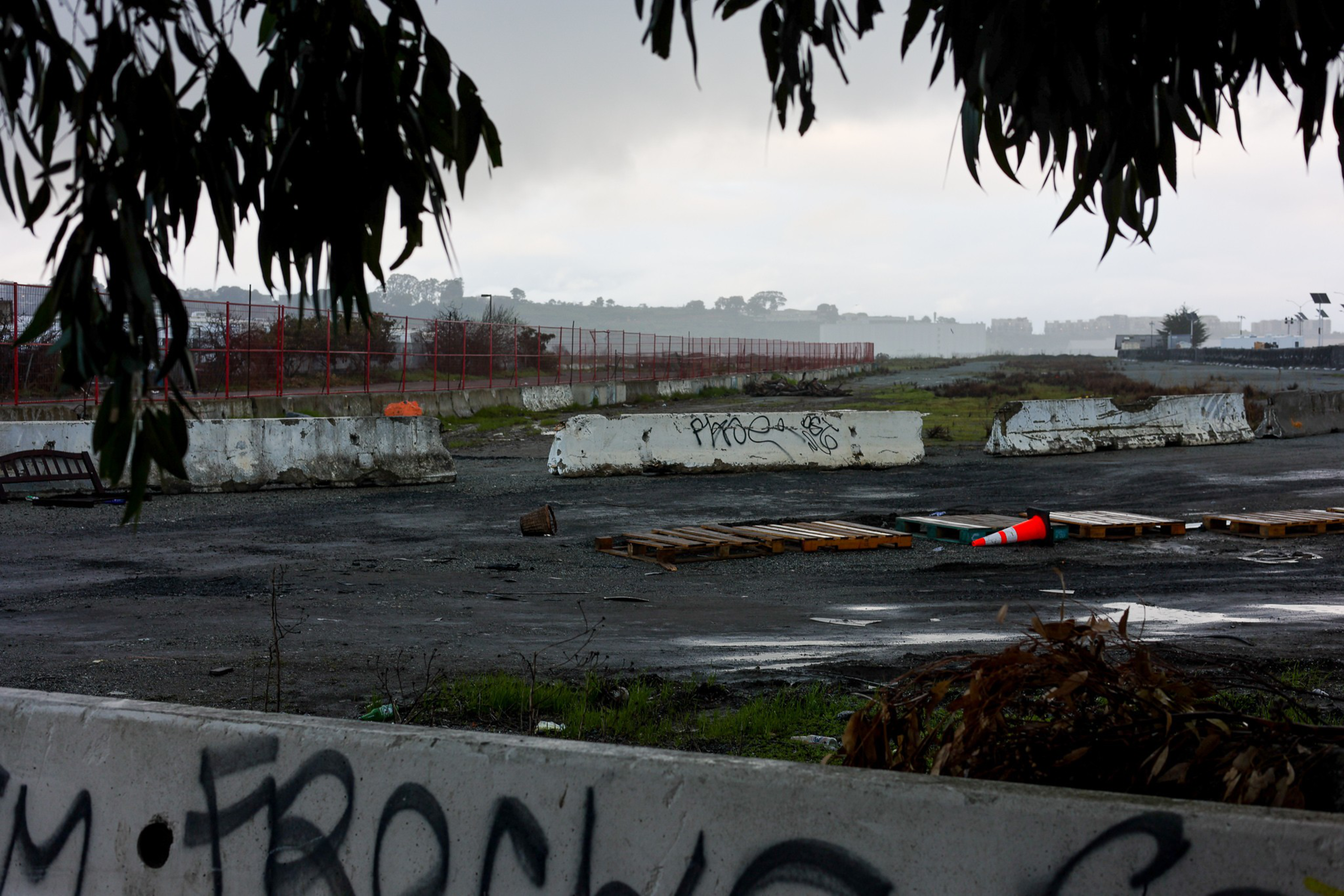 Concrete barriers with graffiti, wooden pallets, and an orange traffic cone are scattered on a wet, empty lot under overcast skies.