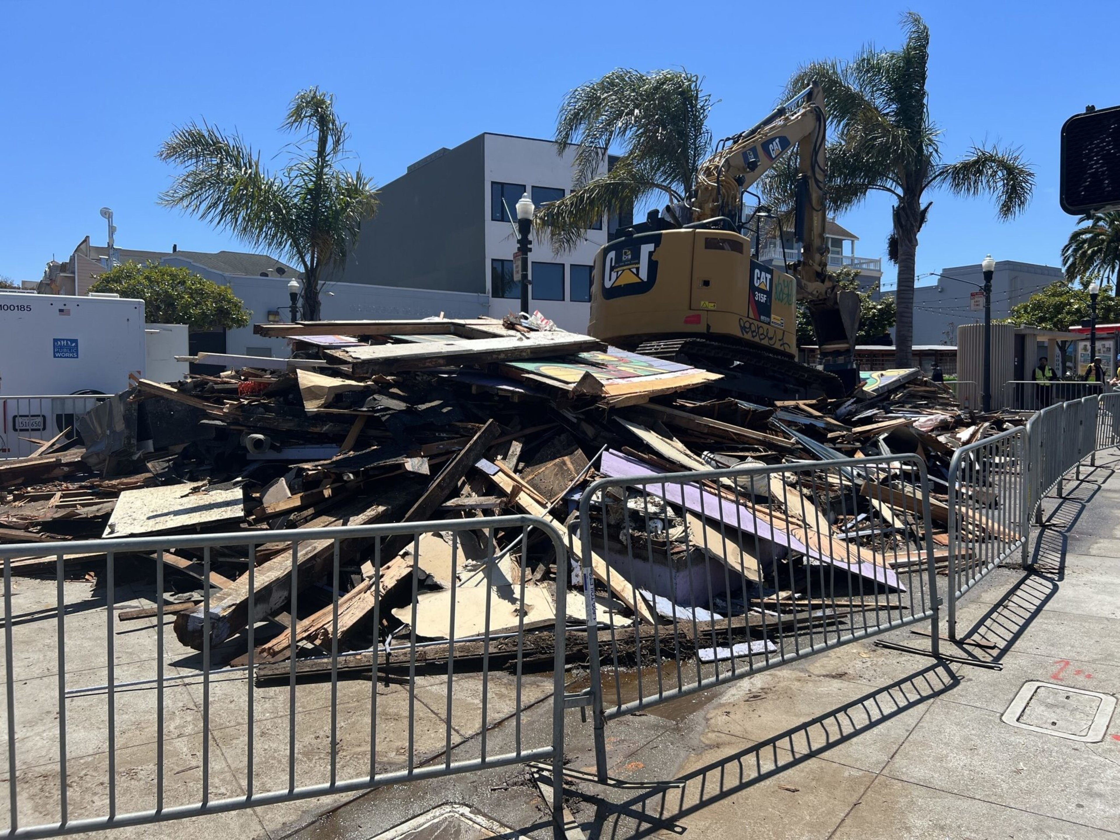 A pile of wooden debris is fenced off on a sunny street, with a yellow CAT excavator and palm trees in the background.