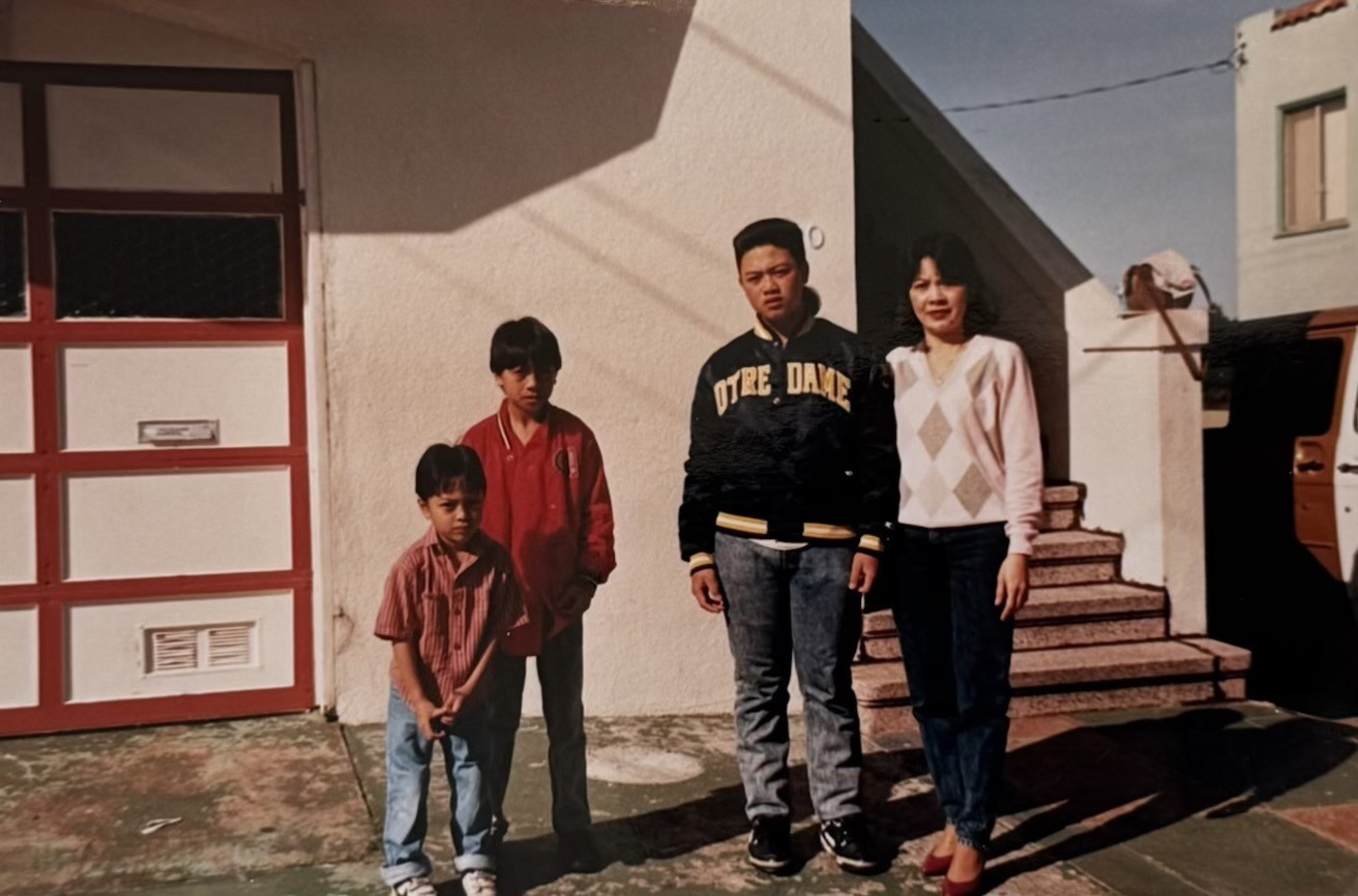 A woman and three boys stand outside. One wears a &quot;Notre Dame&quot; jacket, another a red jacket, and the smallest has a striped shirt. They're near a staircase and garage.