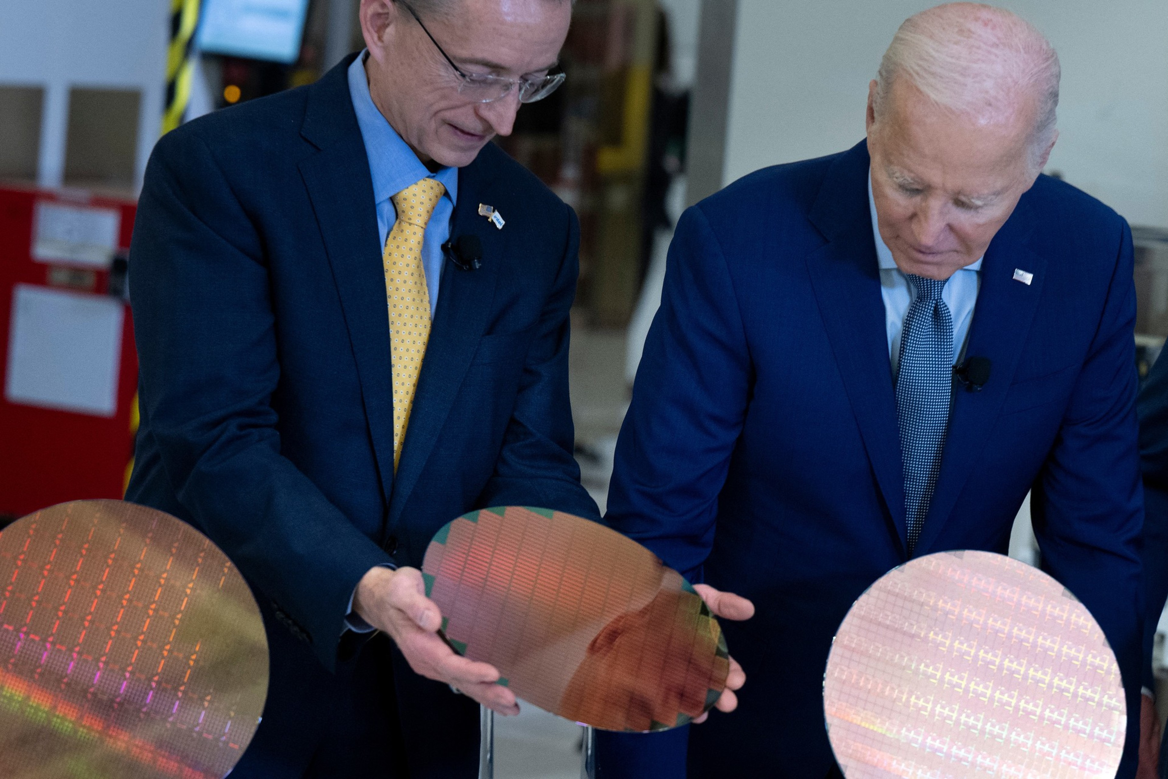 Two men in suits examine reflective silicon wafers, holding them carefully. They are engaged in discussion, appearing in a high-tech or industrial setting.