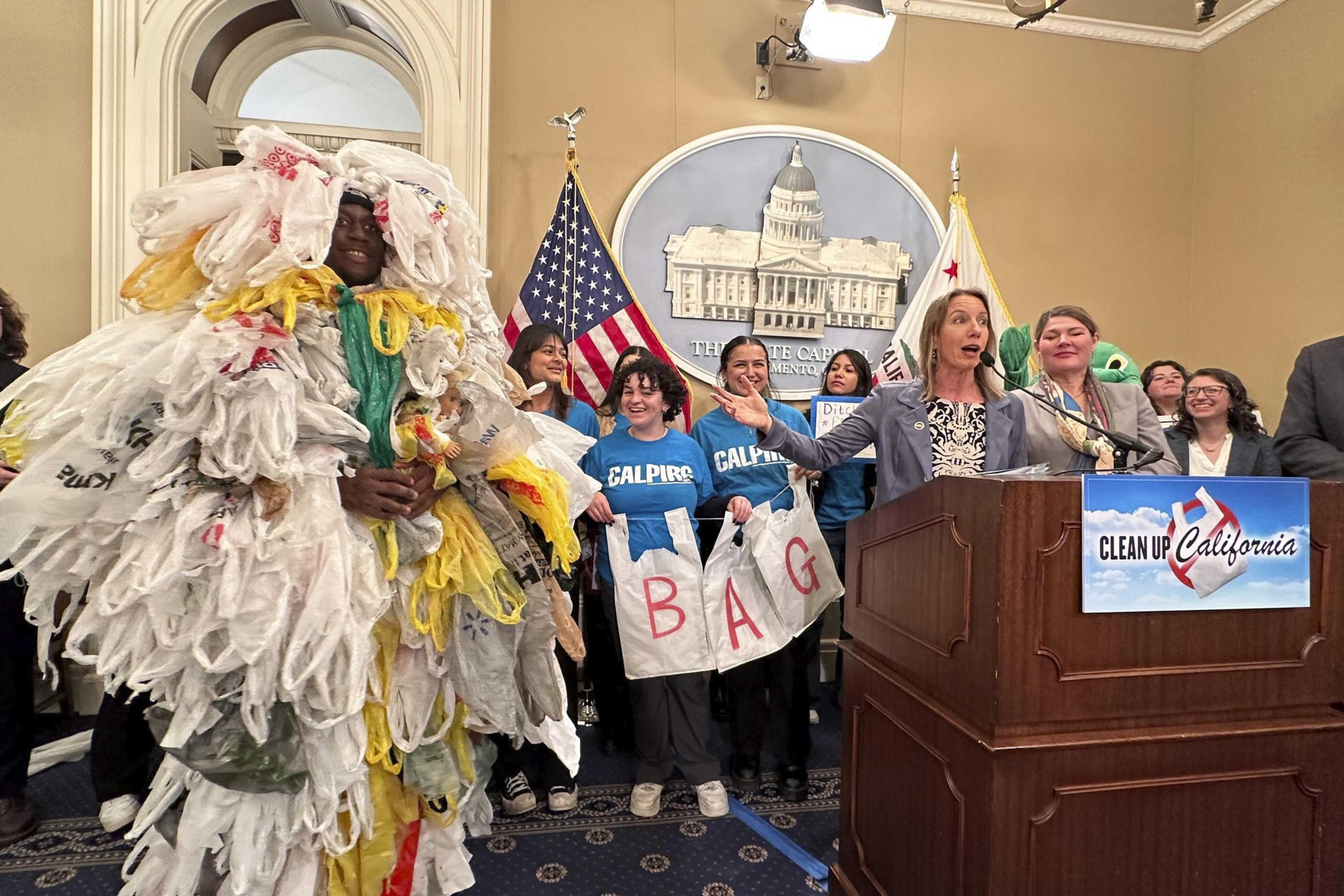A person covered in white yellow and gray plastic bags stands next to group with woman speaking in front of a podium with sign that reads clean up california