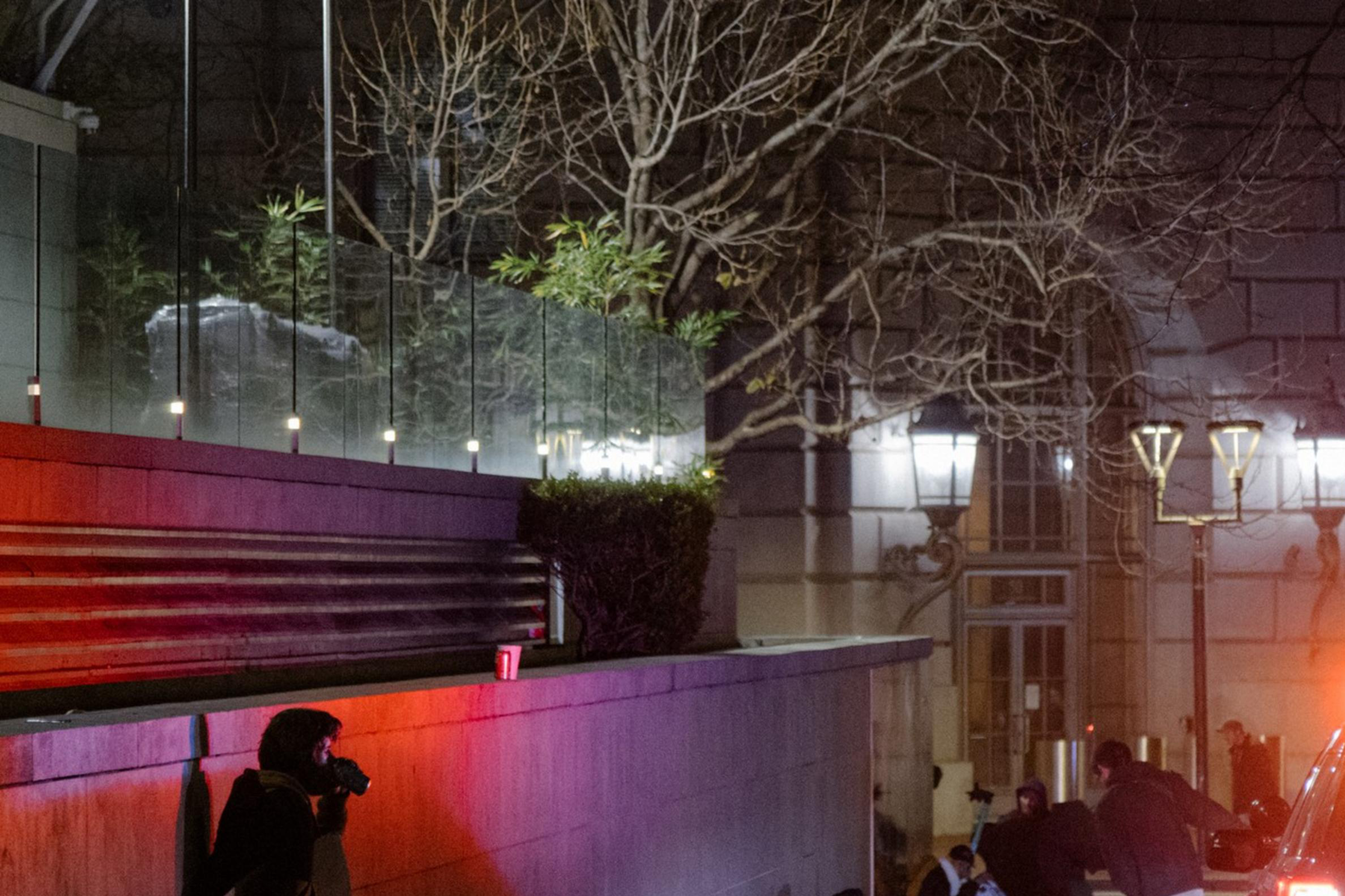 A police car with flashing lights is parked on a dimly lit street beside a building. People are gathered on the sidewalk, and a person is leaning against a wall.