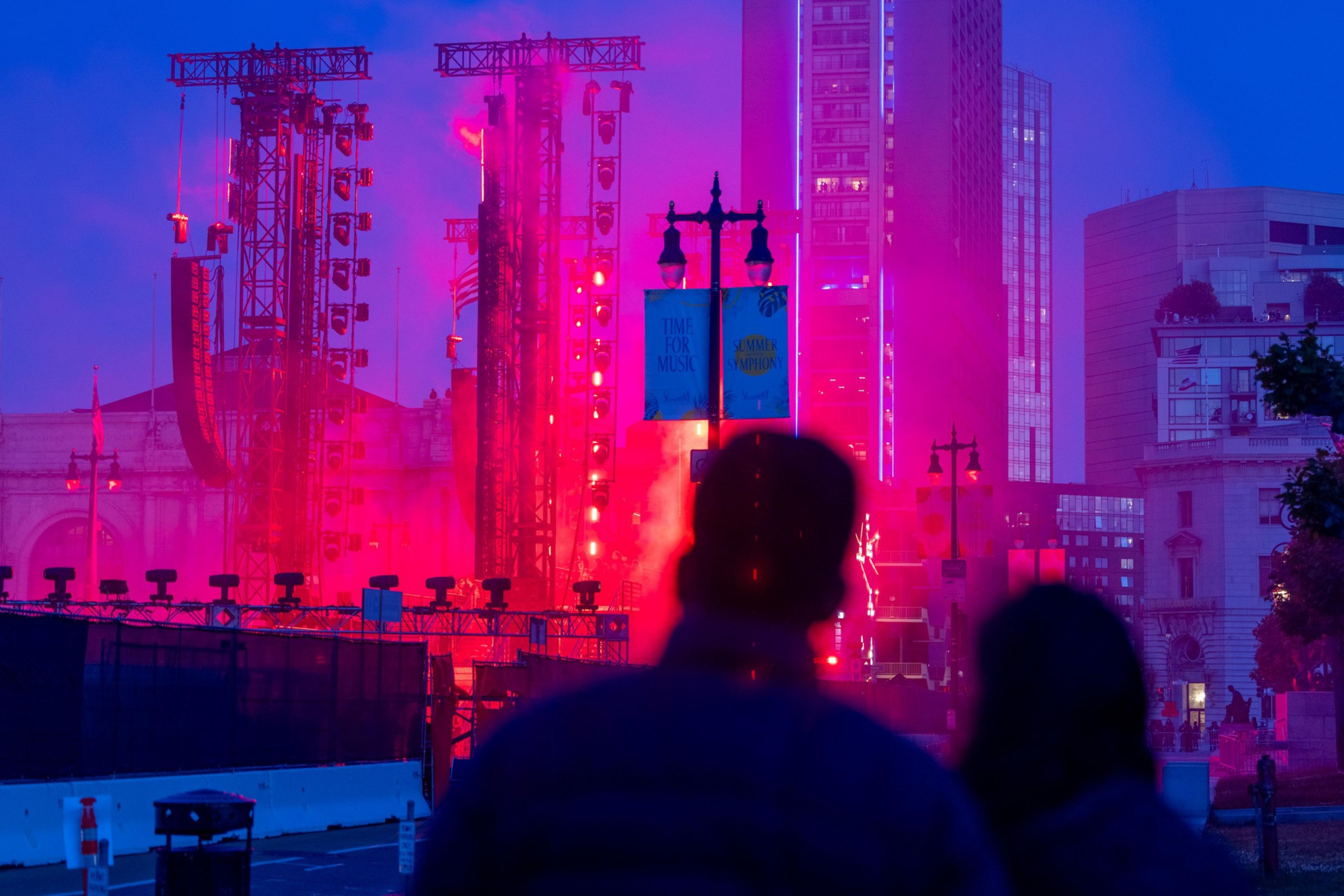 Two figures face a vibrant urban scene with bright red lights, stage equipment, and tall buildings at dusk. Banners read &quot;TIME FOR MUSIC&quot; and &quot;SUMMER SYMPHONY.&quot;