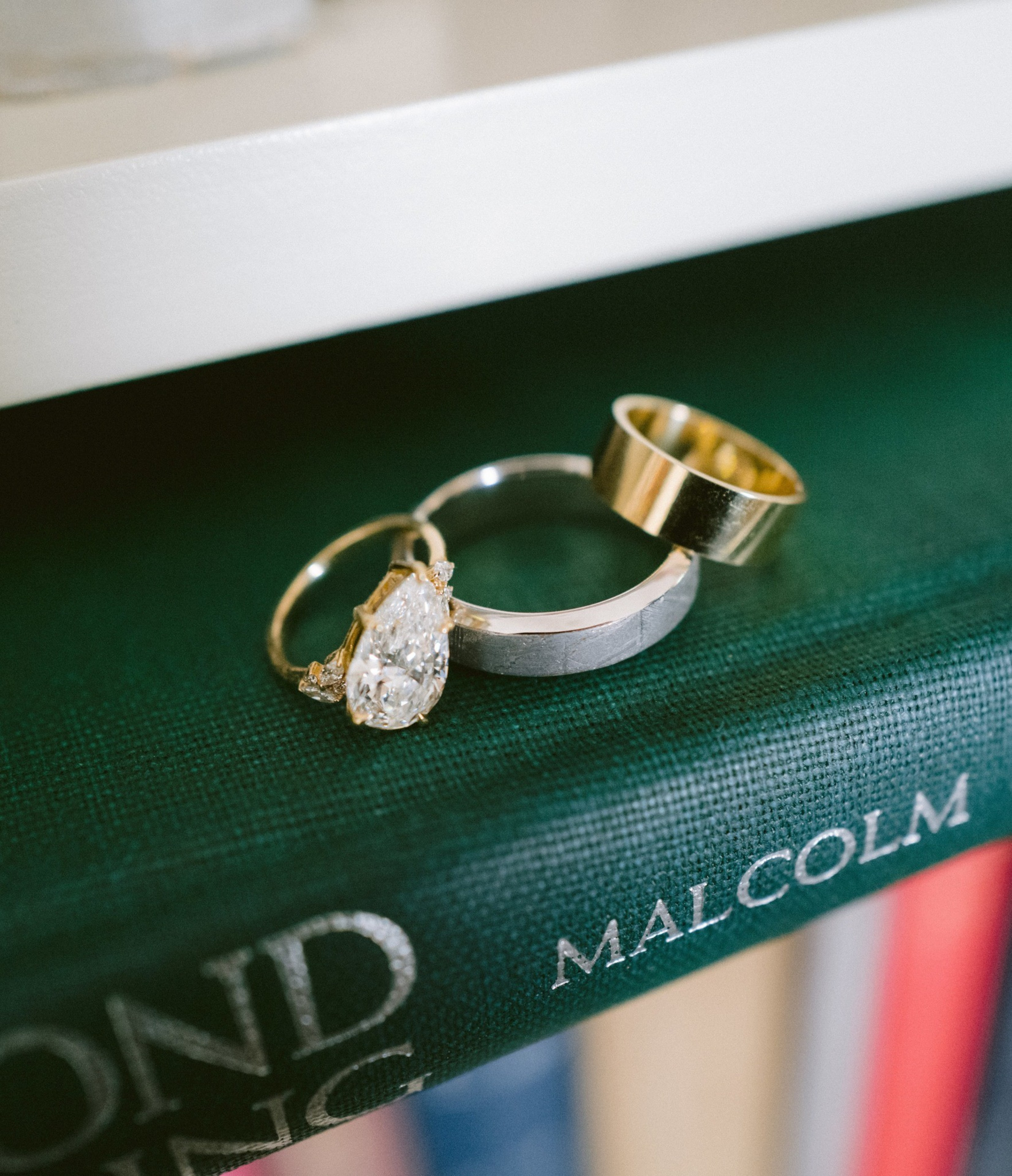 Three rings—a pear-shaped diamond engagement ring, a silver band, and a gold band—rest on the spine of a green book titled “Malcolm.”