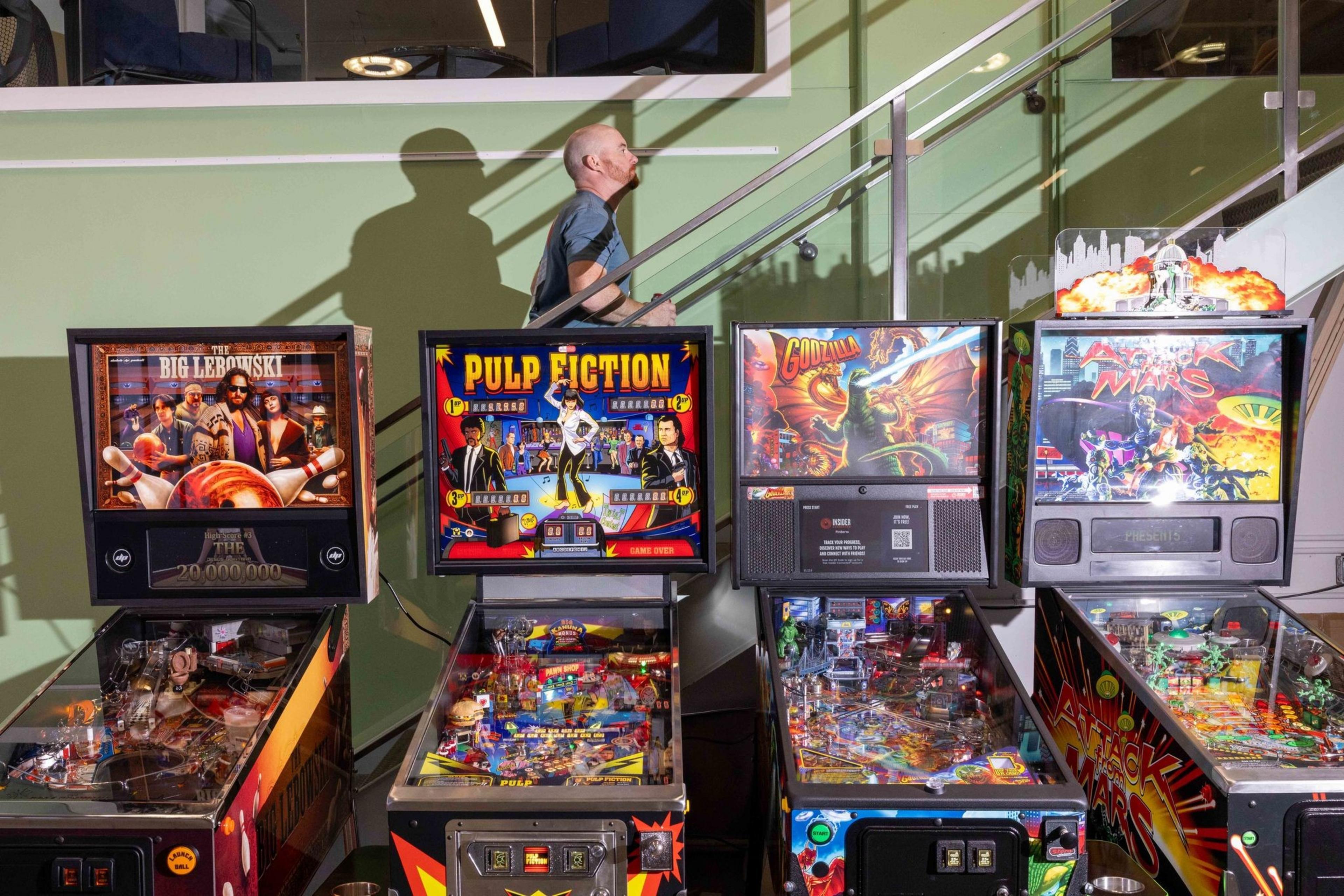 Four pinball machines with vibrant themed artwork stand in front of a green wall. A person is seen walking up a staircase in the background.