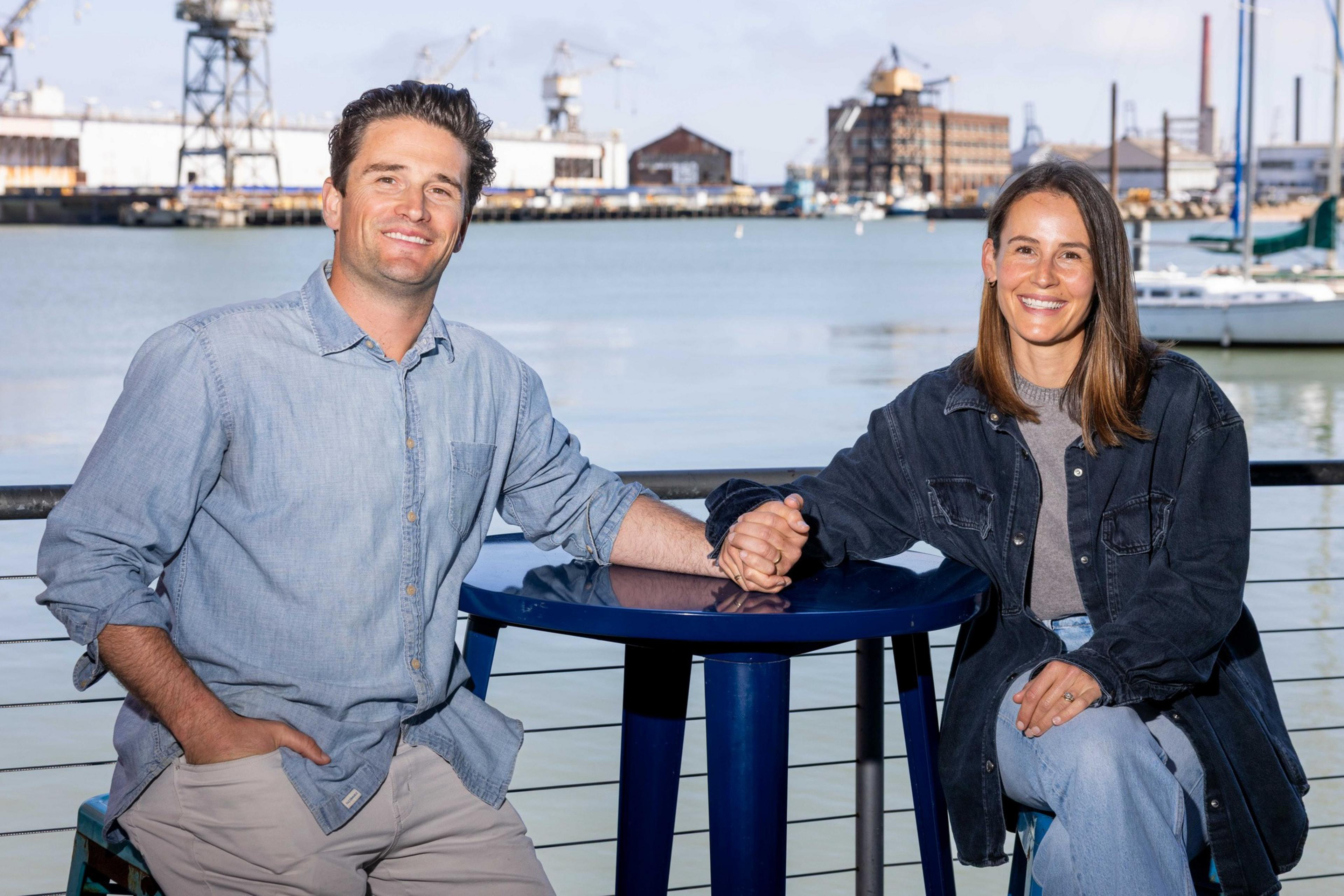 A smiling couple holds hands across a blue table, sitting by a railing with a harbor and industrial buildings in the background.