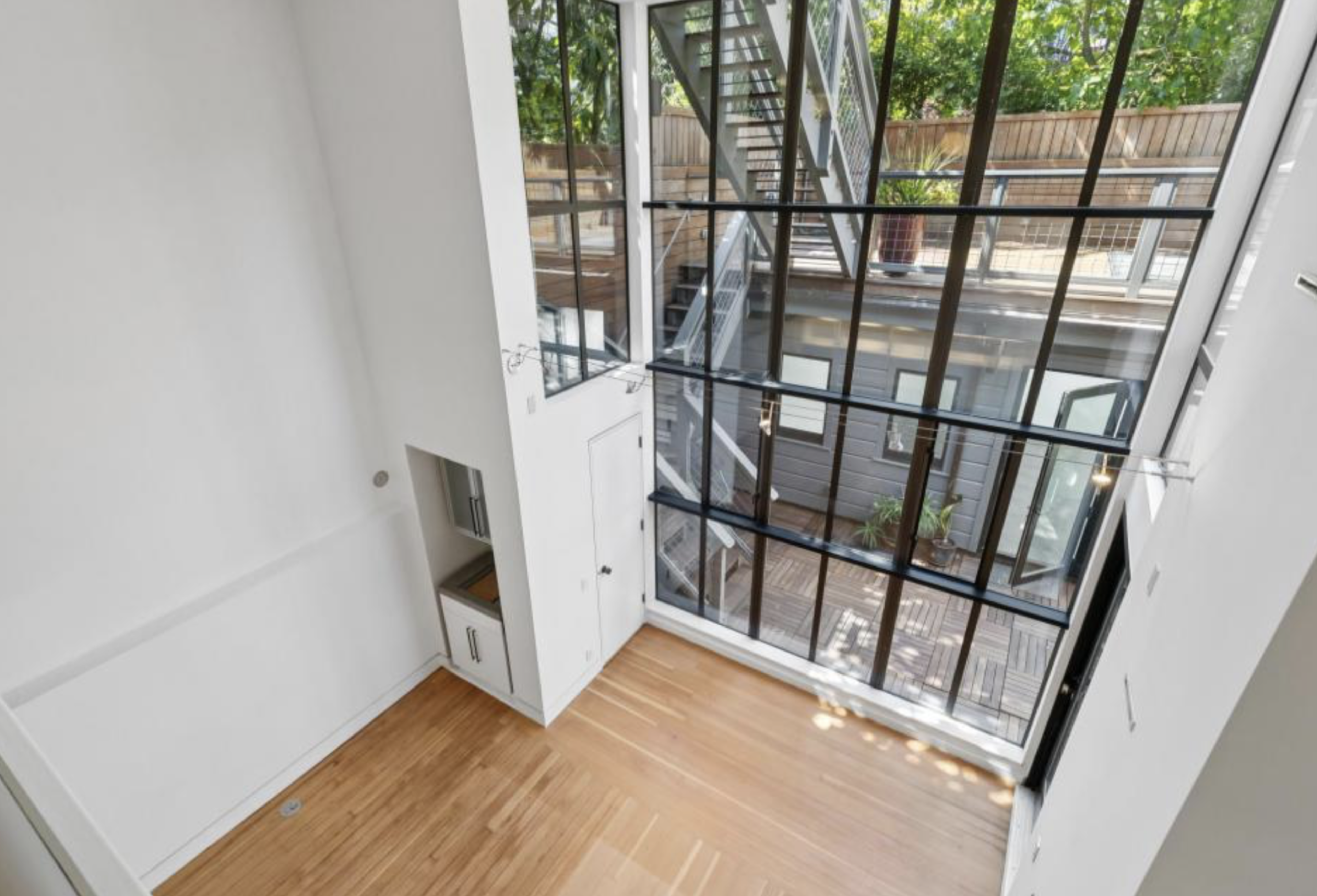 A bright, empty room with high ceilings, light wood floors, white walls, and a tall black-framed glass window overlooking an outdoor deck with stairs.