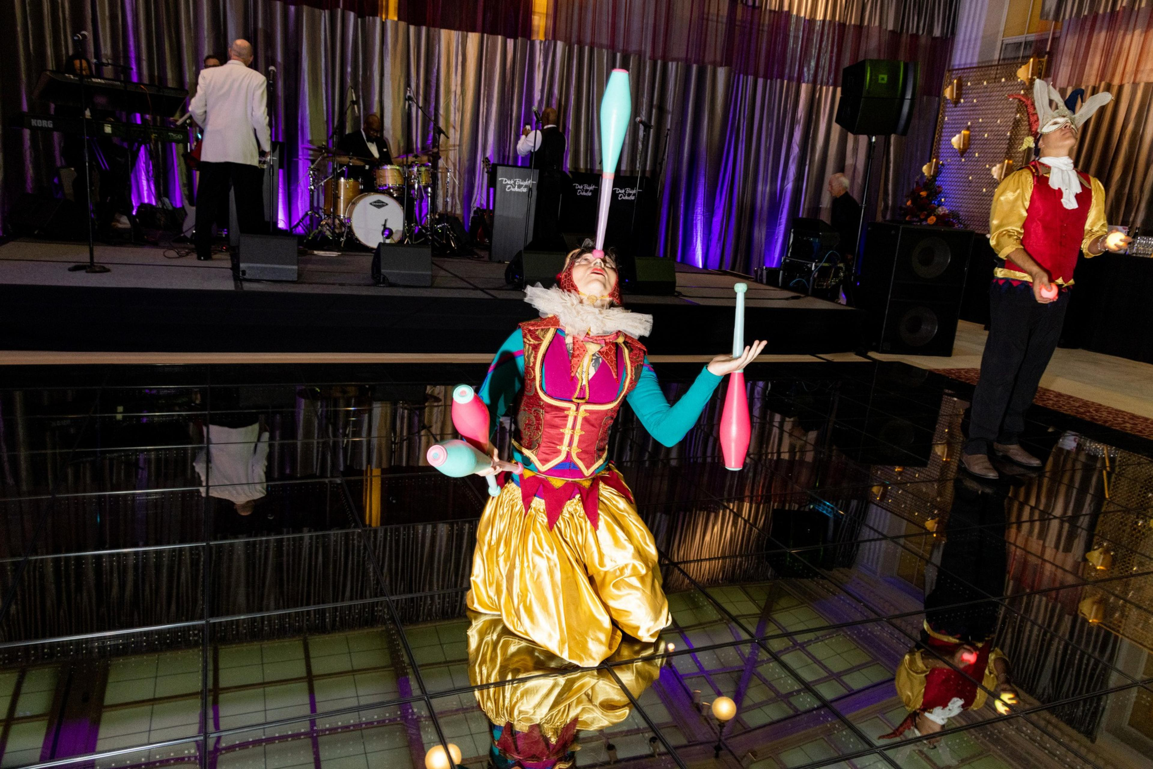 A performer dressed in a colorful jester costume kneels while juggling five clubs, with a stage and another performer in a similar costume nearby.