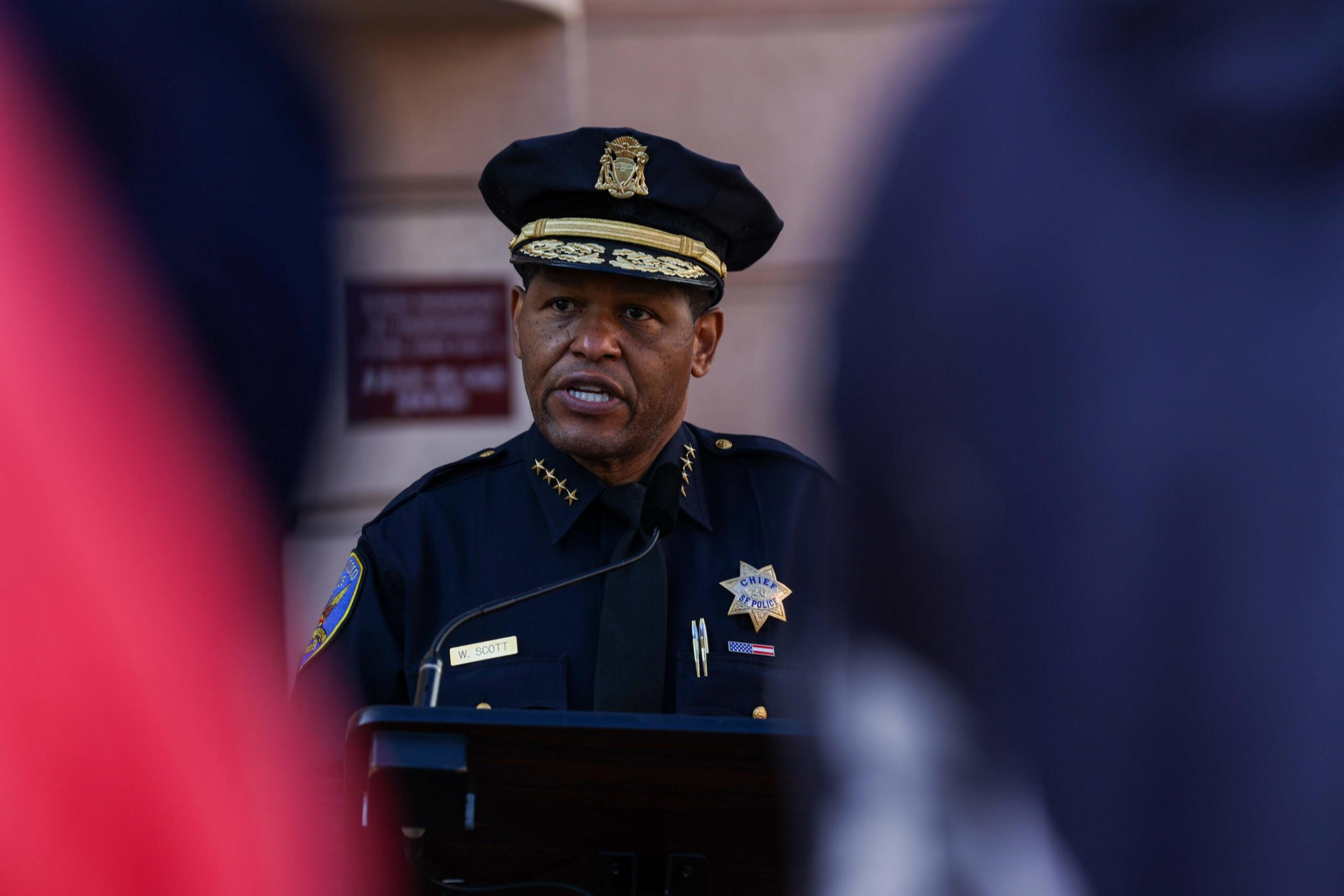 A police chief in uniform speaks at a podium, framed by blurred figures in the foreground, with a building wall behind him.