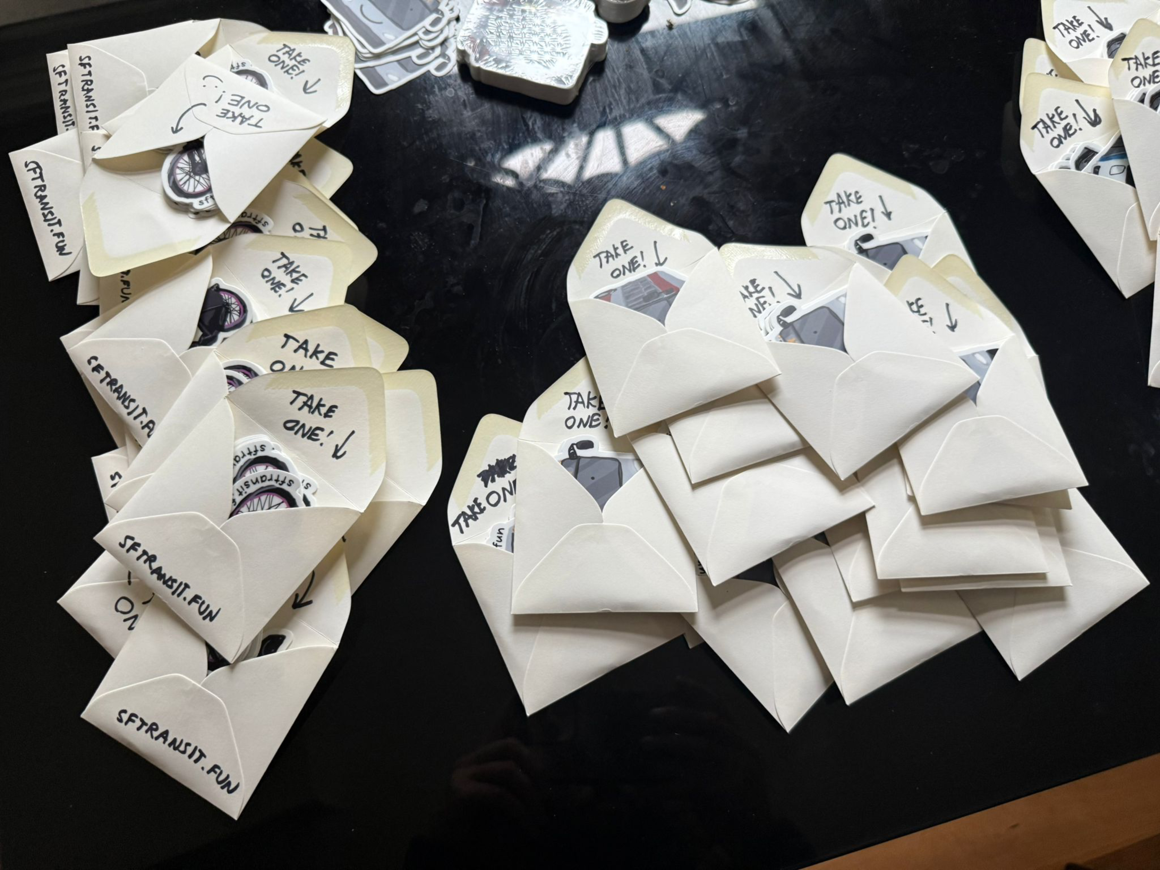 The image shows several small envelopes labeled &quot;TAKE ONE!&quot; lying on a black surface. Many contain round pins with a design featuring a wheel and text.