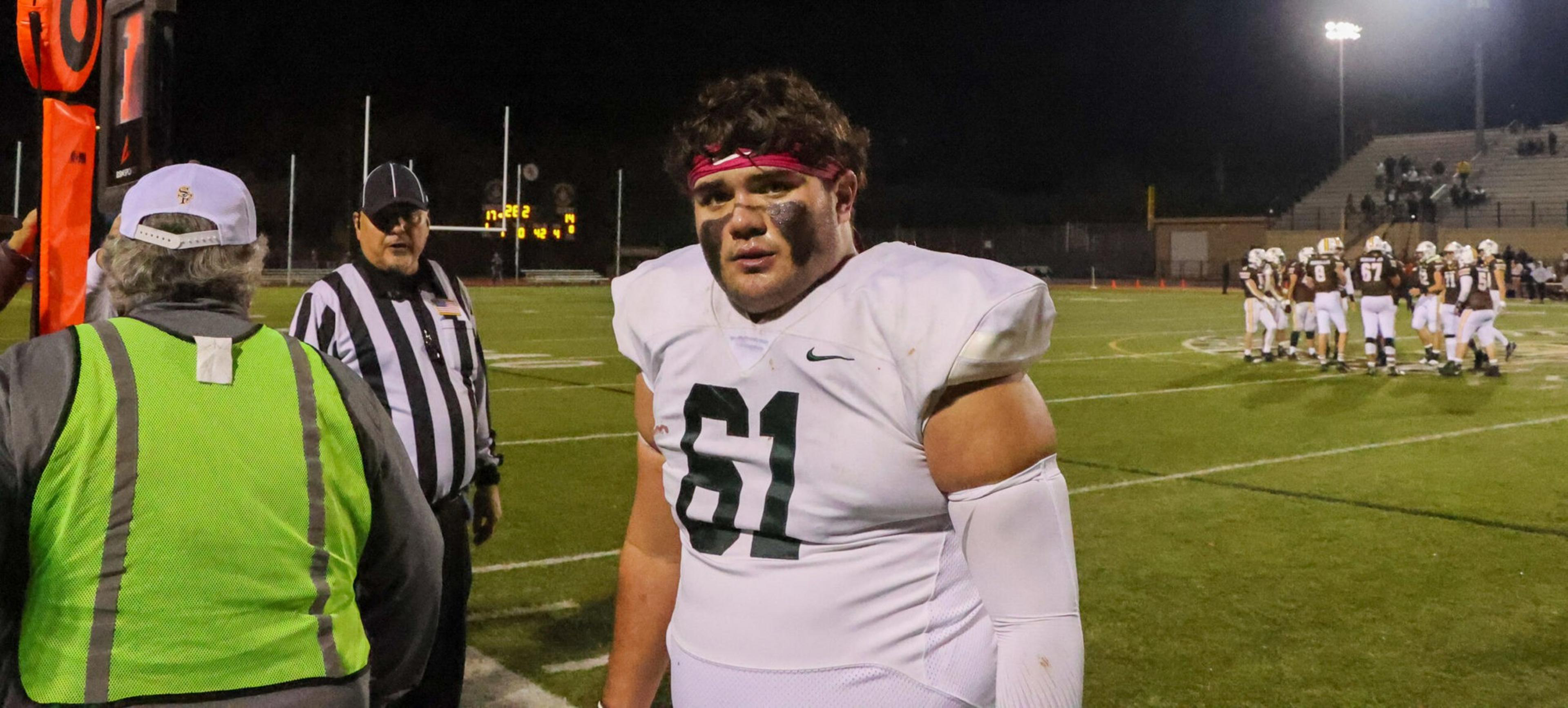 A football player wearing a white jersey with number 61 and eye black stands on the field near a referee and official during a night game.
