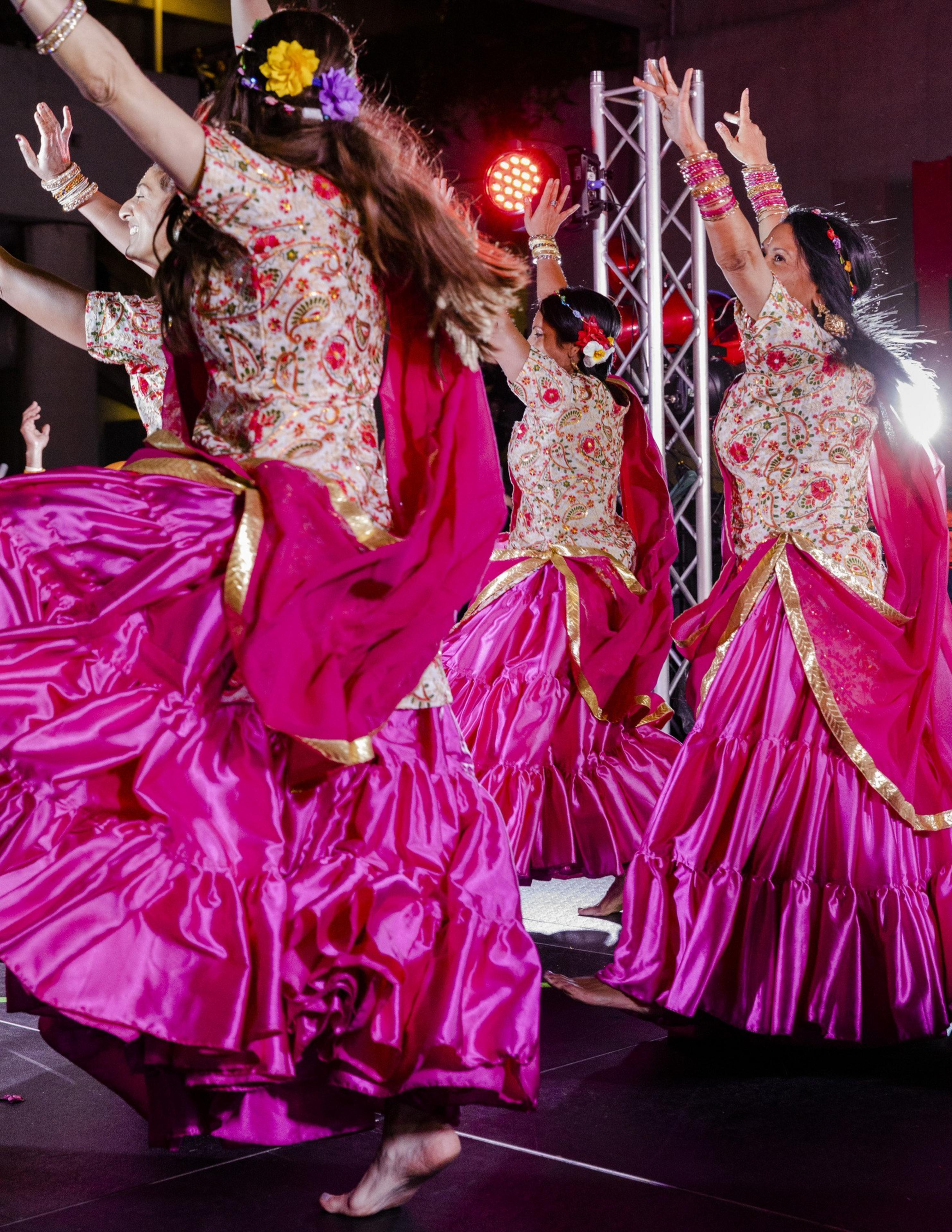 A group of dancers in vibrant pink, gold-trimmed skirts and floral-patterned tops perform energetically, their hands raised, against a backdrop of colorful stage lights.
