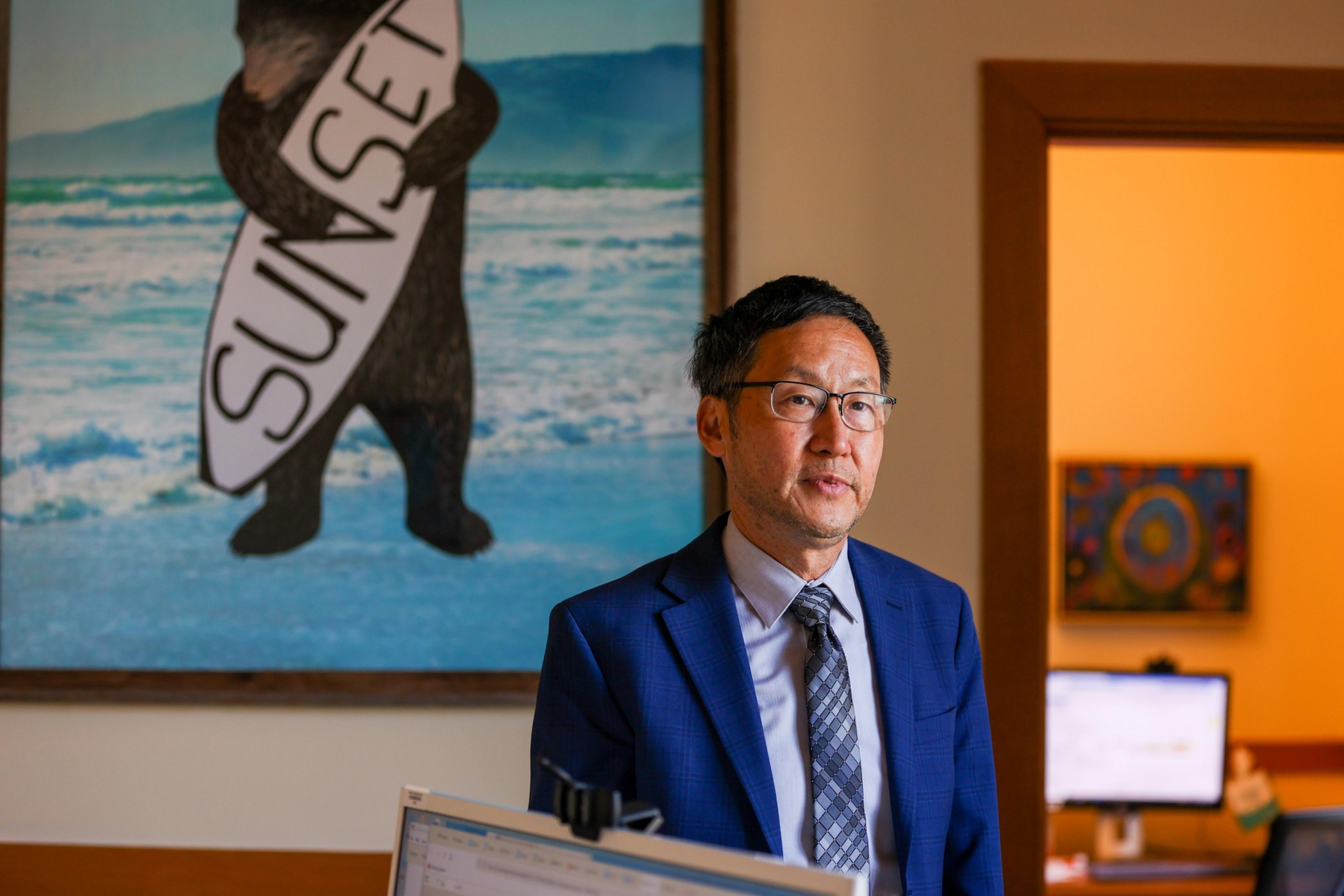 A man in glasses and a blue suit stands indoors near a computer, with a painting of a bear holding a "SUNSET" surfboard and a brightly lit room behind him.